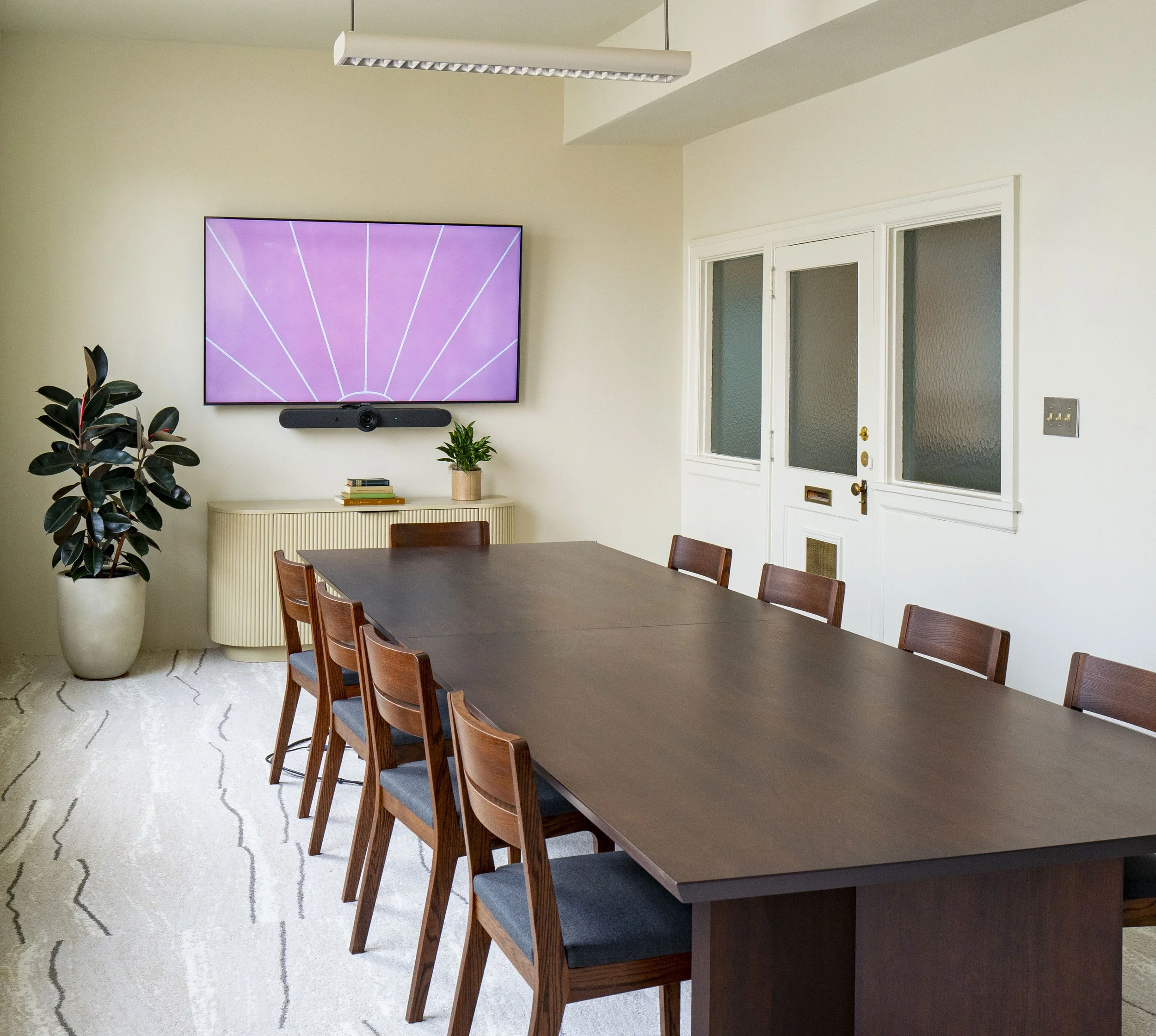 Modern dining room with a rectangular dark wood table and six matching chairs with navy blue cushions. A large flat-screen TV is mounted on the wall above a cream-colored console table with a few books and a potted plant. To the left, a tall potted plant with dark green leaves is visible. The room has white walls, a textured white door with frosted glass panels, and a patterned white area rug on a light hardwood floor. There is a white modern ceiling light fixture.