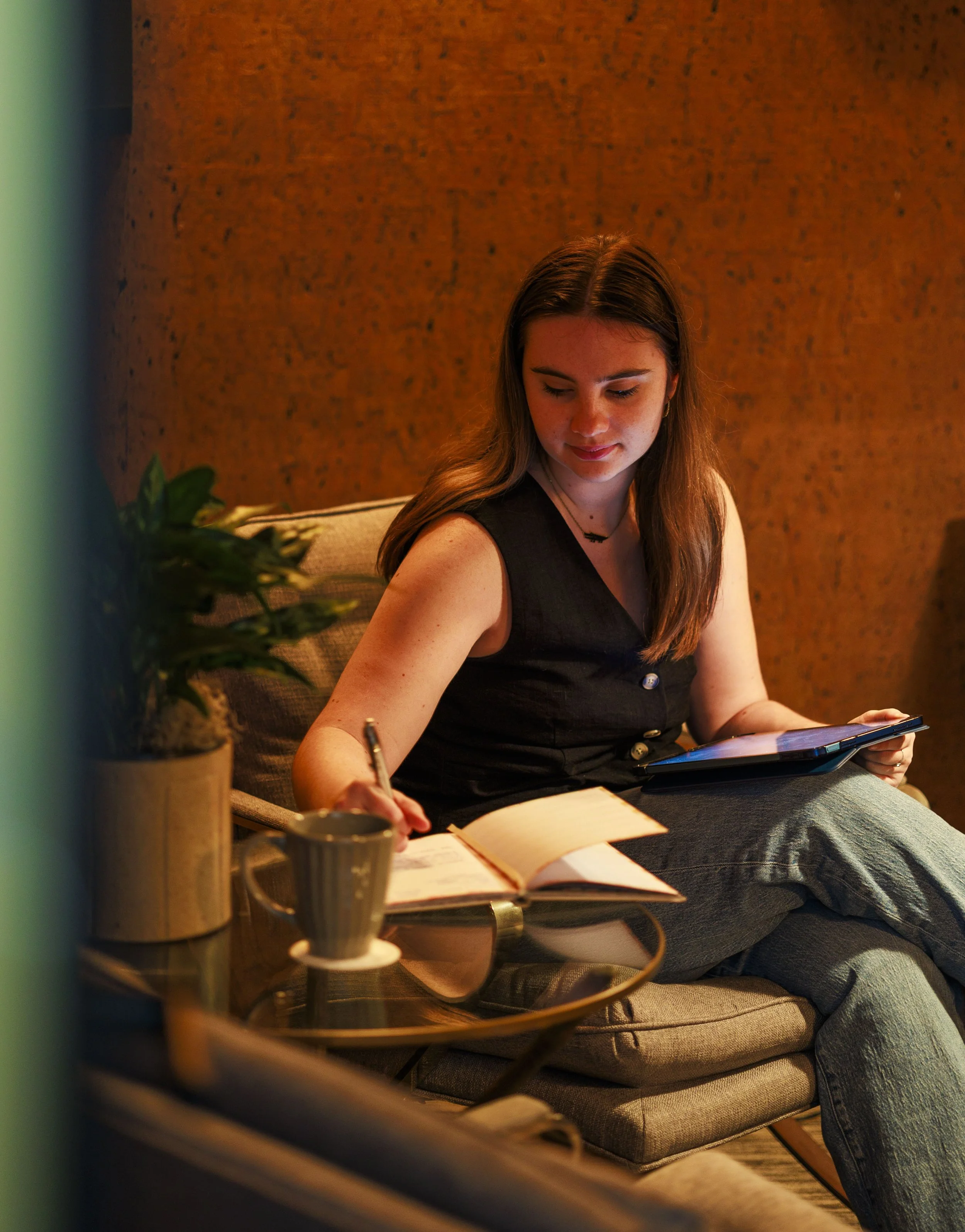 A young woman sitting on a beige sofa, looking at a tablet in her right hand, with an open notebook and pen on her lap, in a warmly lit living room with a wooden wall background, a small potted plant, and a coffee cup on a round wooden table in the foreground.