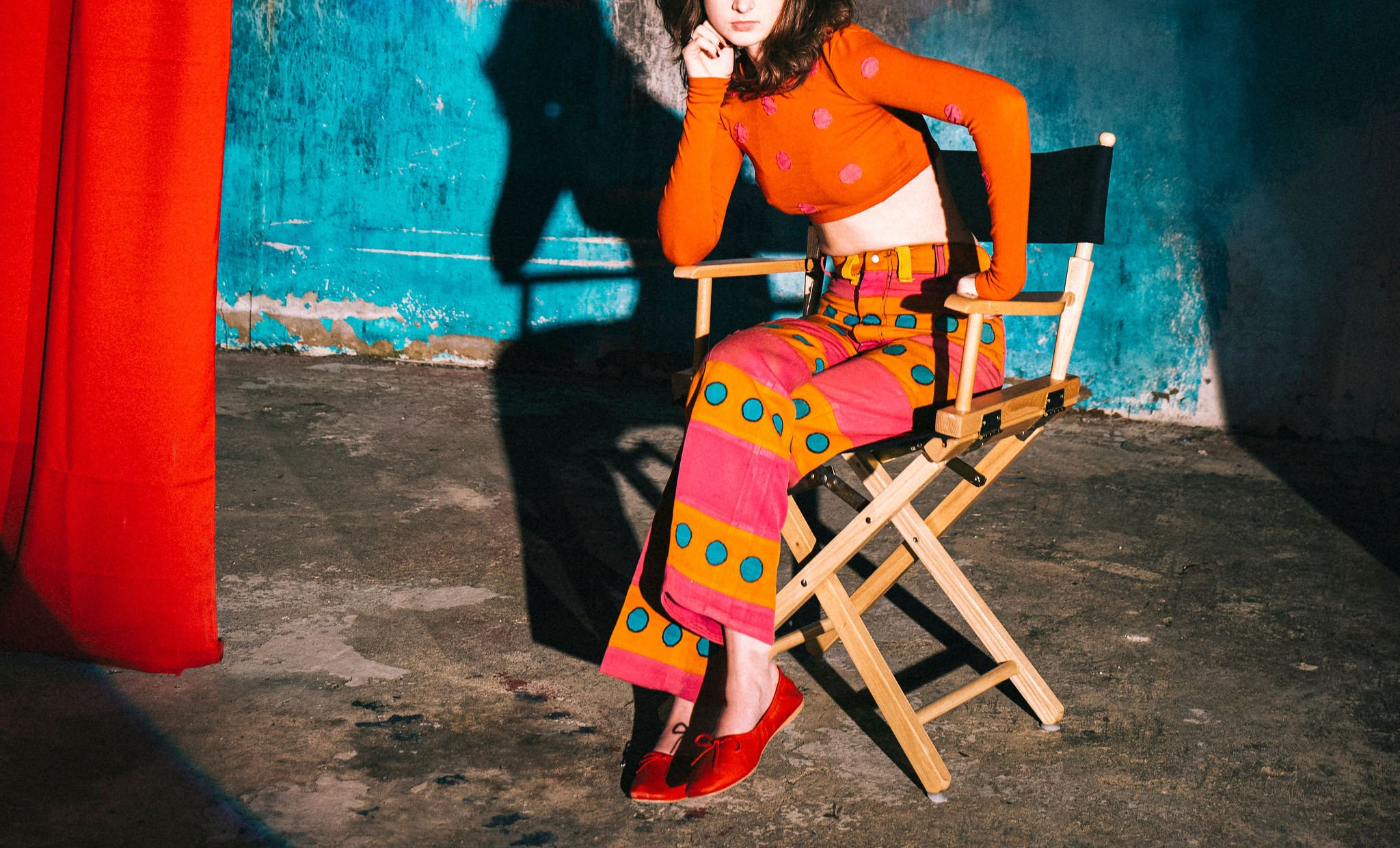 A woman sitting on a wooden chair with a blue wall and a red curtain nearby. She is wearing a colorful outfit with an orange long-sleeve top with pink polka dots, patterned pants with pink, yellow, and blue circles, and red shoes. She has dark curly 