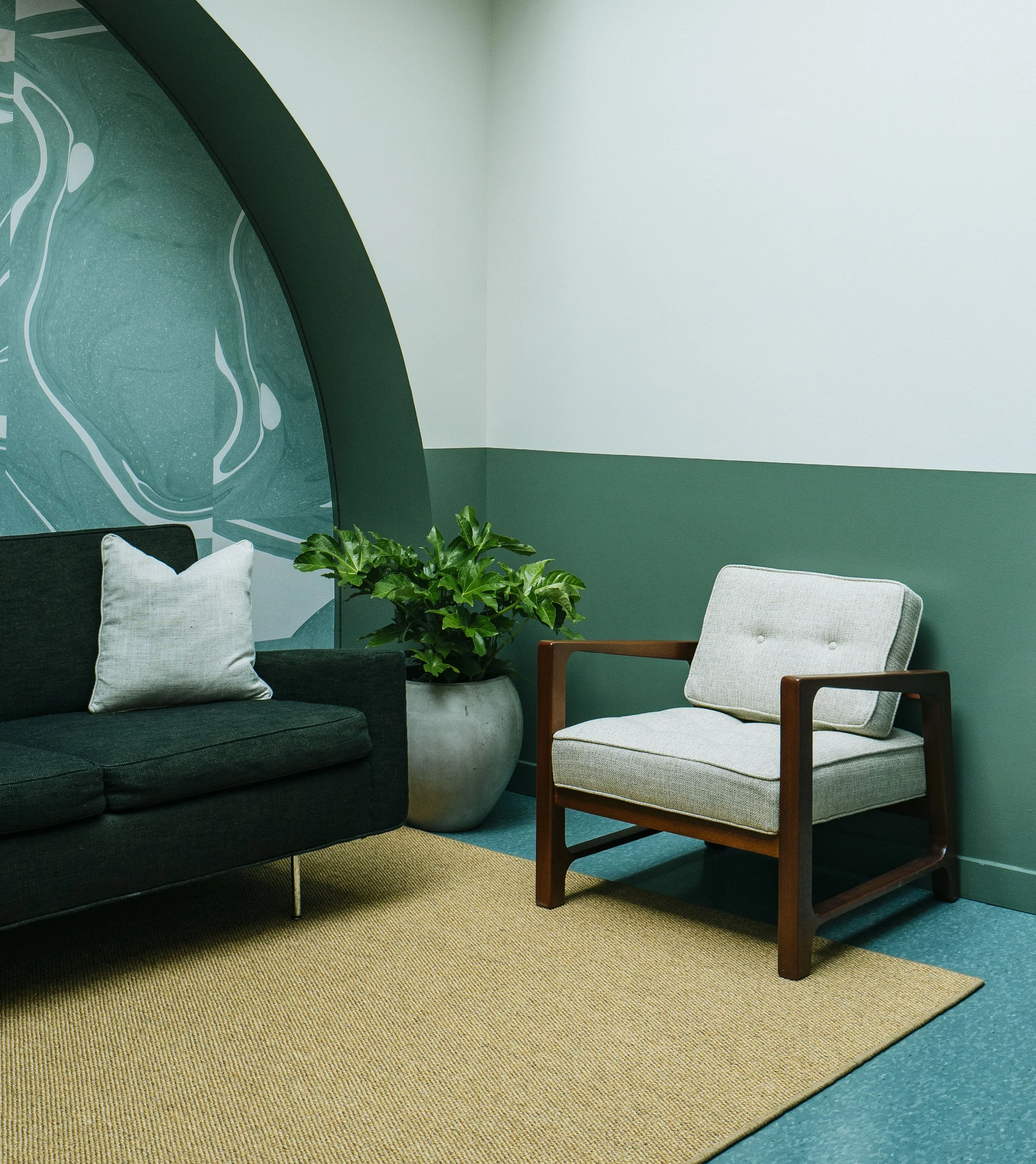 A seating area with a black sofa with a white pillow, a beige armchair with a wooden frame, a large potted leafy plant, a beige rug, and a green wall with a decorative glass panel and a corner of a white wall.
