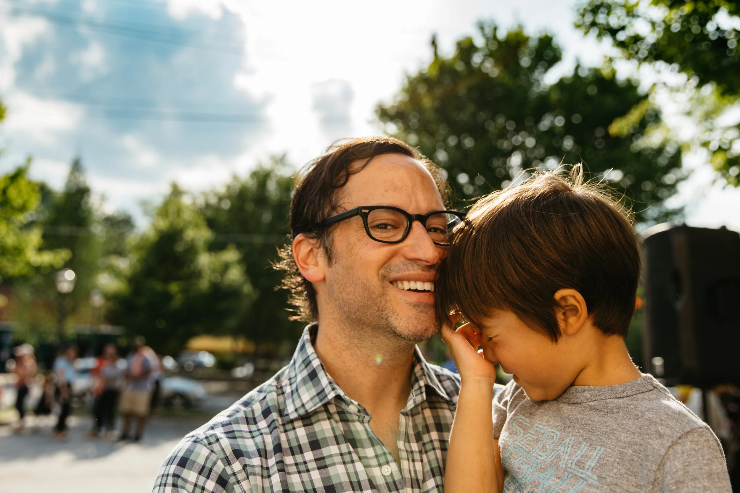 A man with glasses smiles as a child, likely his son, rests his face against his. They are outside in a park or public space on a sunny day, with trees and a crowd of people in the background.