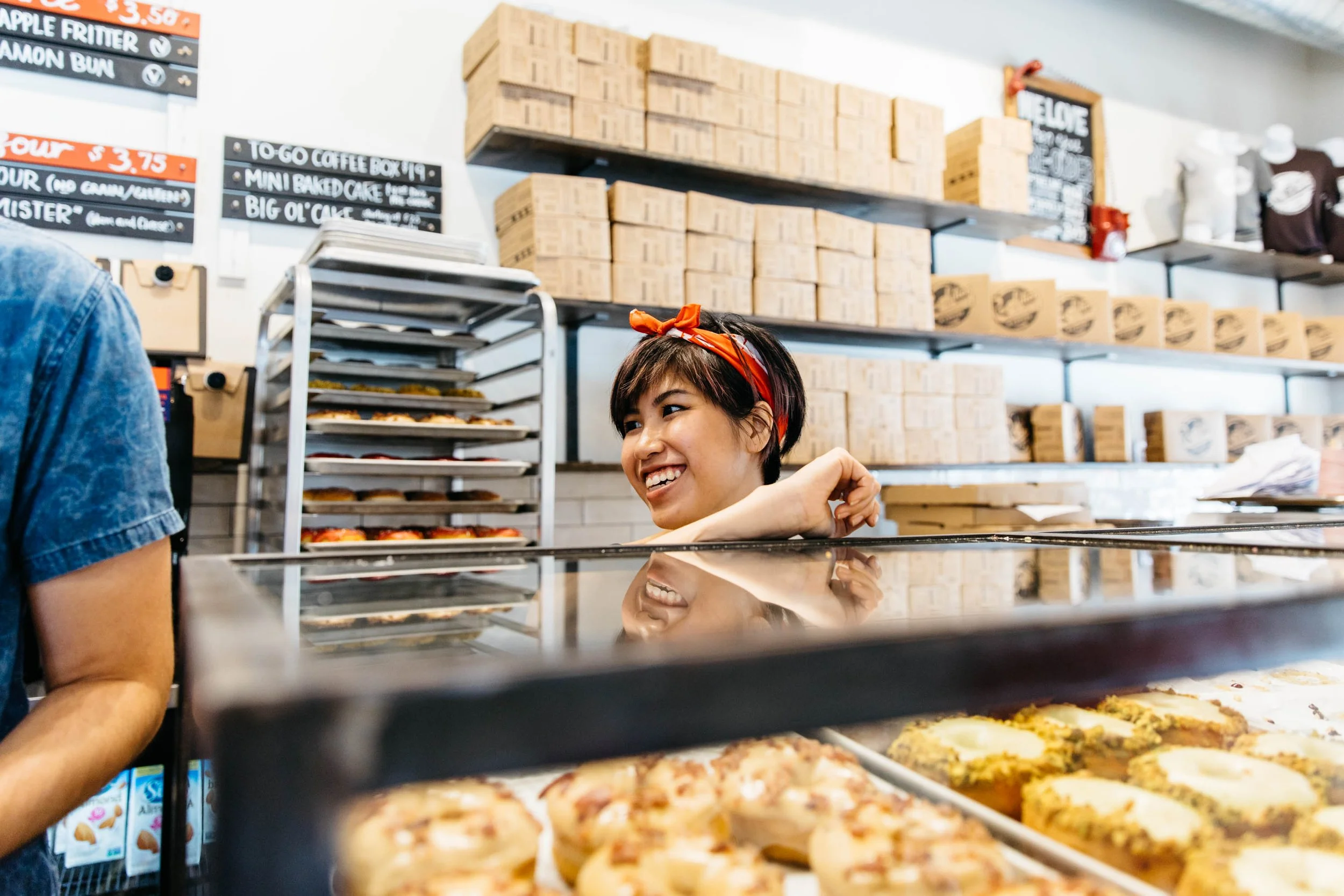 Smiling woman at pizzeria counter with pizza trays in the foreground and menu boards on the wall behind her.
