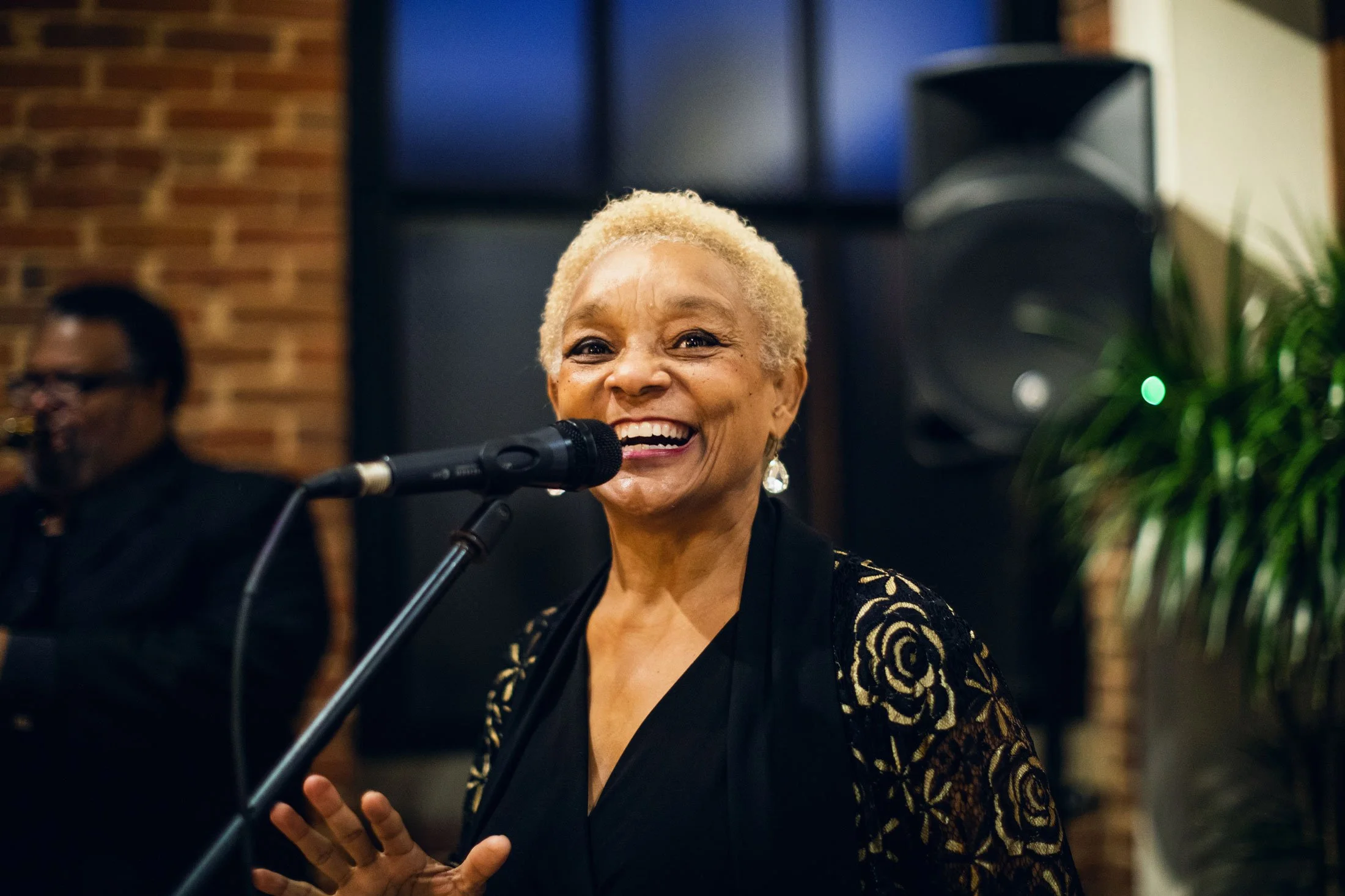 Smiling elderly woman with short blonde hair singing into a microphone at an indoor event, with a man and a speaker visible in the background.