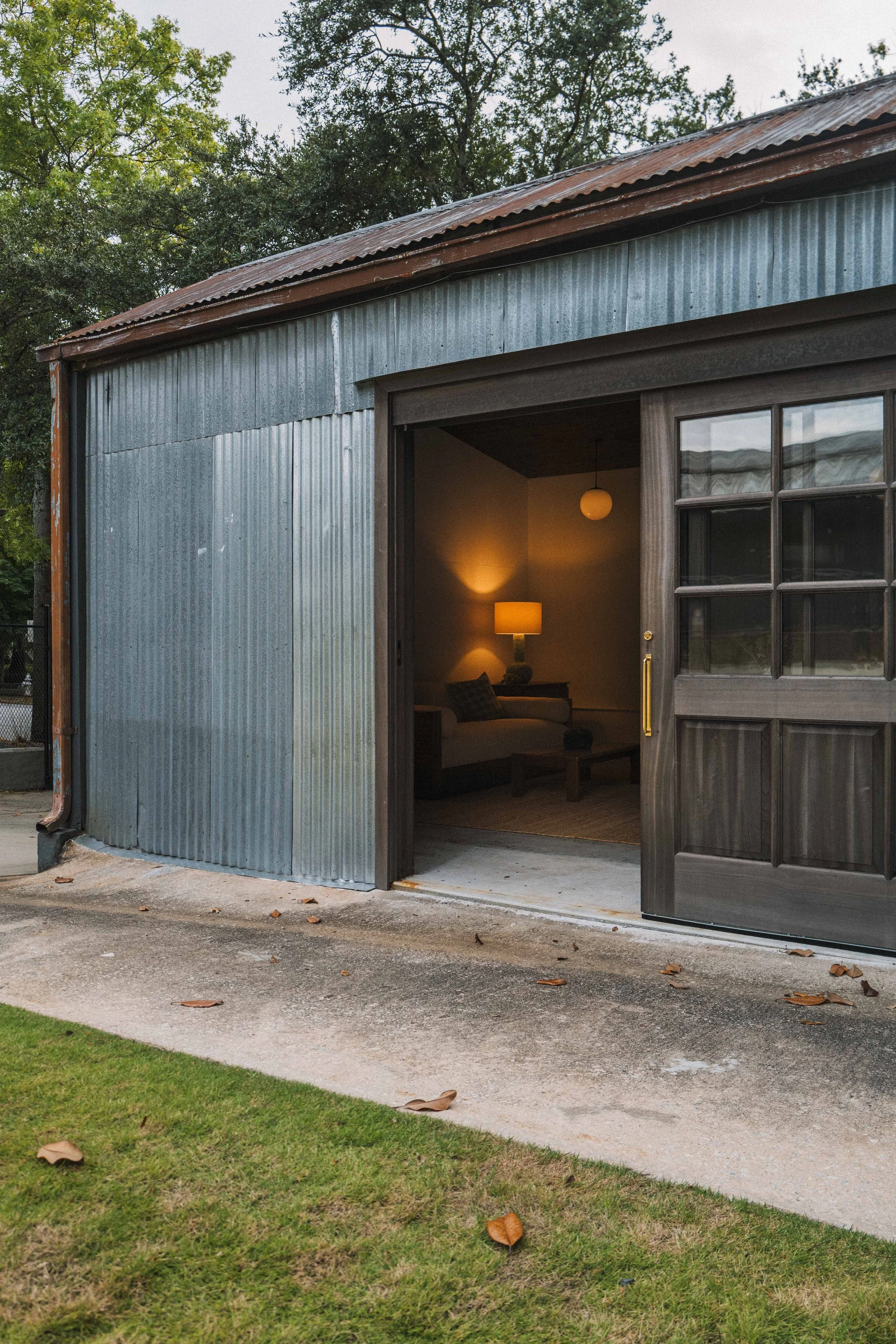 A rustic metal and wood garage with large open sliding door, interior lit warm with a lamp, outside is a grassy area with fallen leaves, and trees in the background.