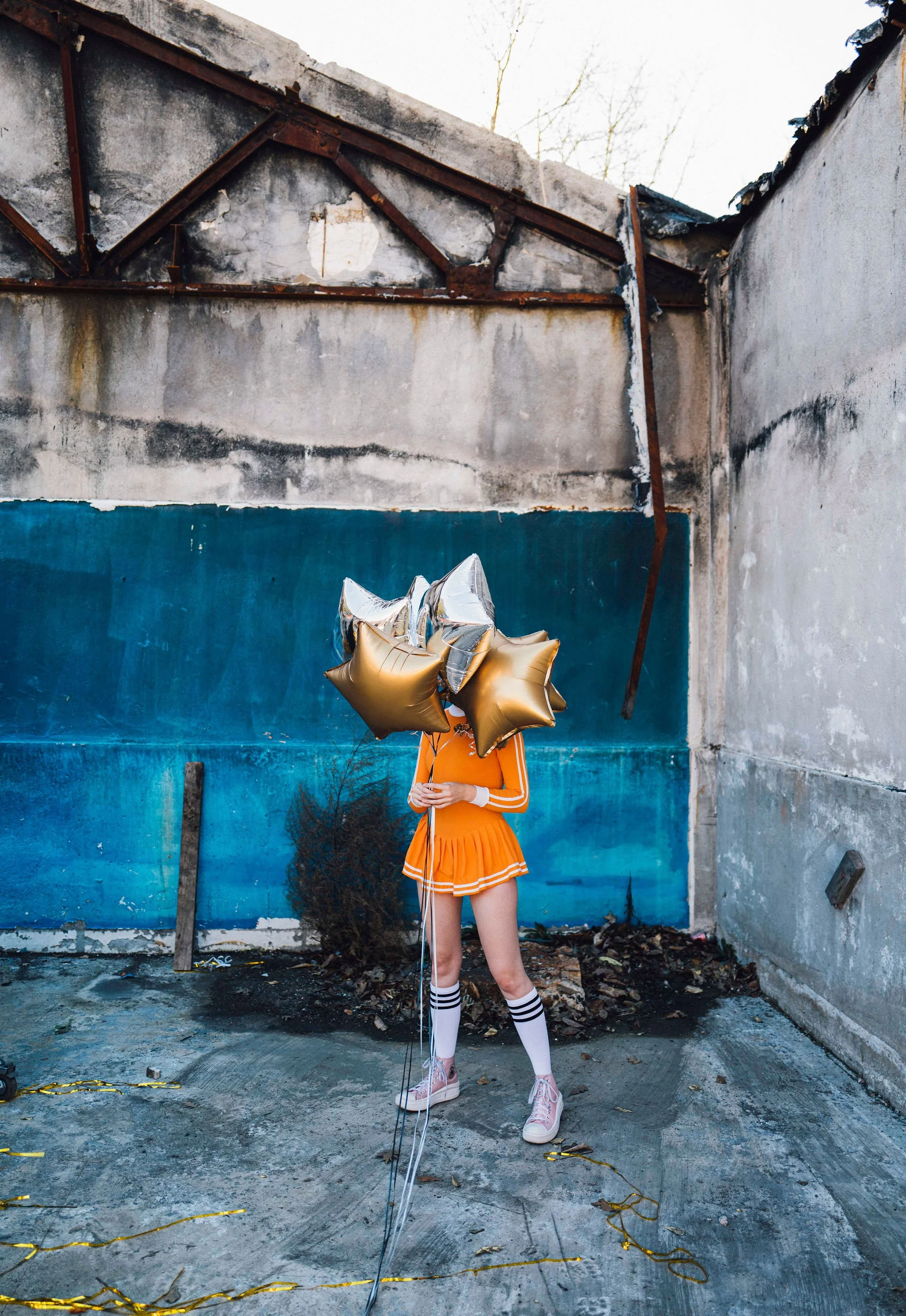 A person in an orange cheerleader outfit, wearing pink sneakers, holding three gold star-shaped balloons, standing in an abandoned setting with weathered walls and debris on the ground.