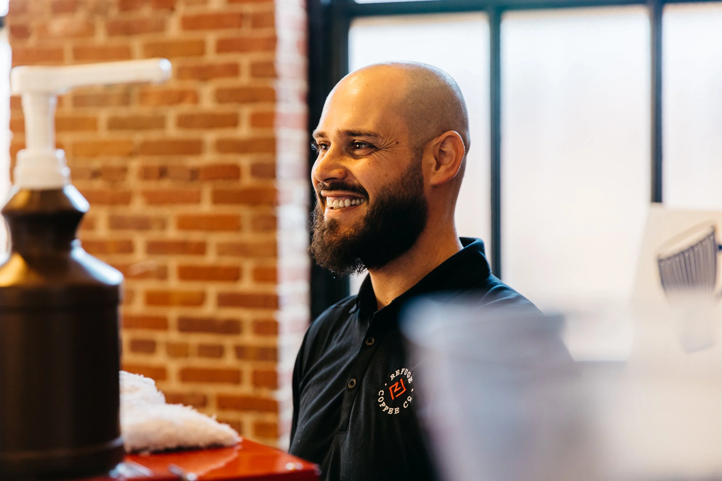A smiling bald man with a beard wears a black polo shirt with a logo, sitting inside a coffee shop with exposed brick walls and large windows in the background.