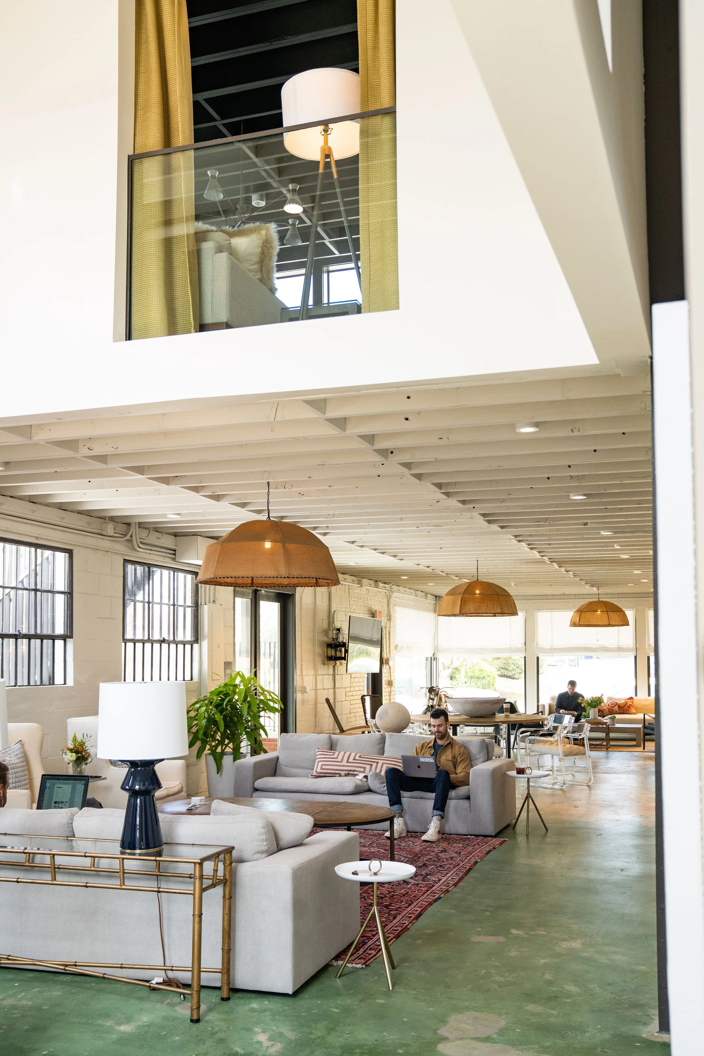 Large open concept living space with multiple seating areas, including sofas and chairs, decorated with plants, lamps, and a patterned rug. Two men are working on laptops in the background, and the room features large windows, hanging pendant lights,