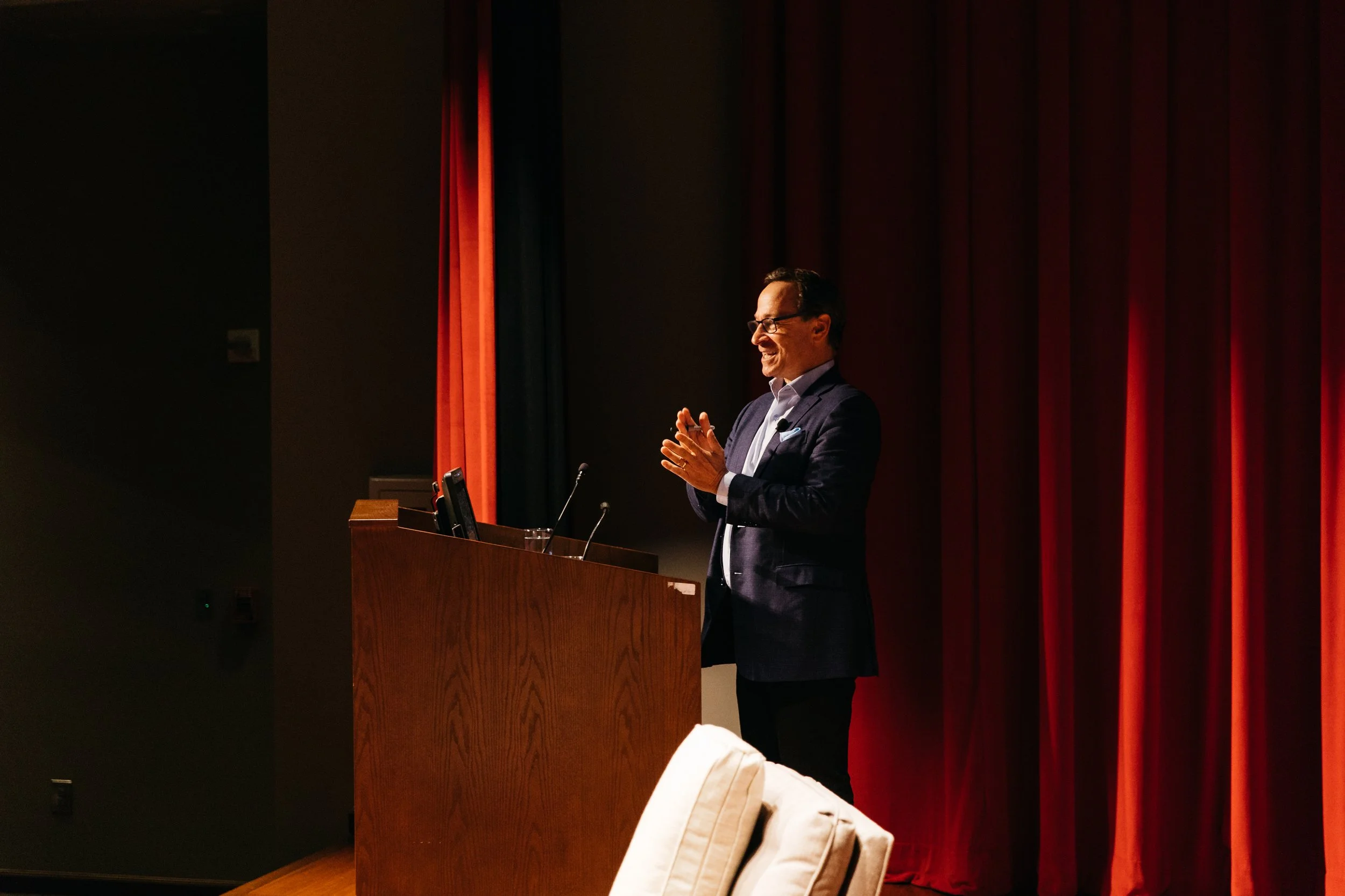 A man in a suit and glasses is standing at a podium, speaking or presenting, with red and black curtains in the background.