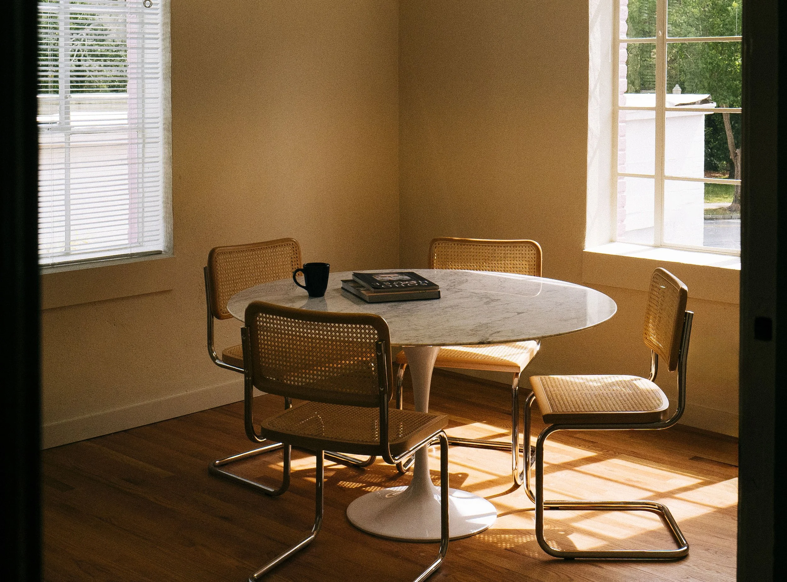 A dining area with a round marble table, four chairs with caned seats and backs, a black mug, and two magazines on the table. There are two large windows with white blinds and a view of green trees outside, allowing natural light to fill the room.