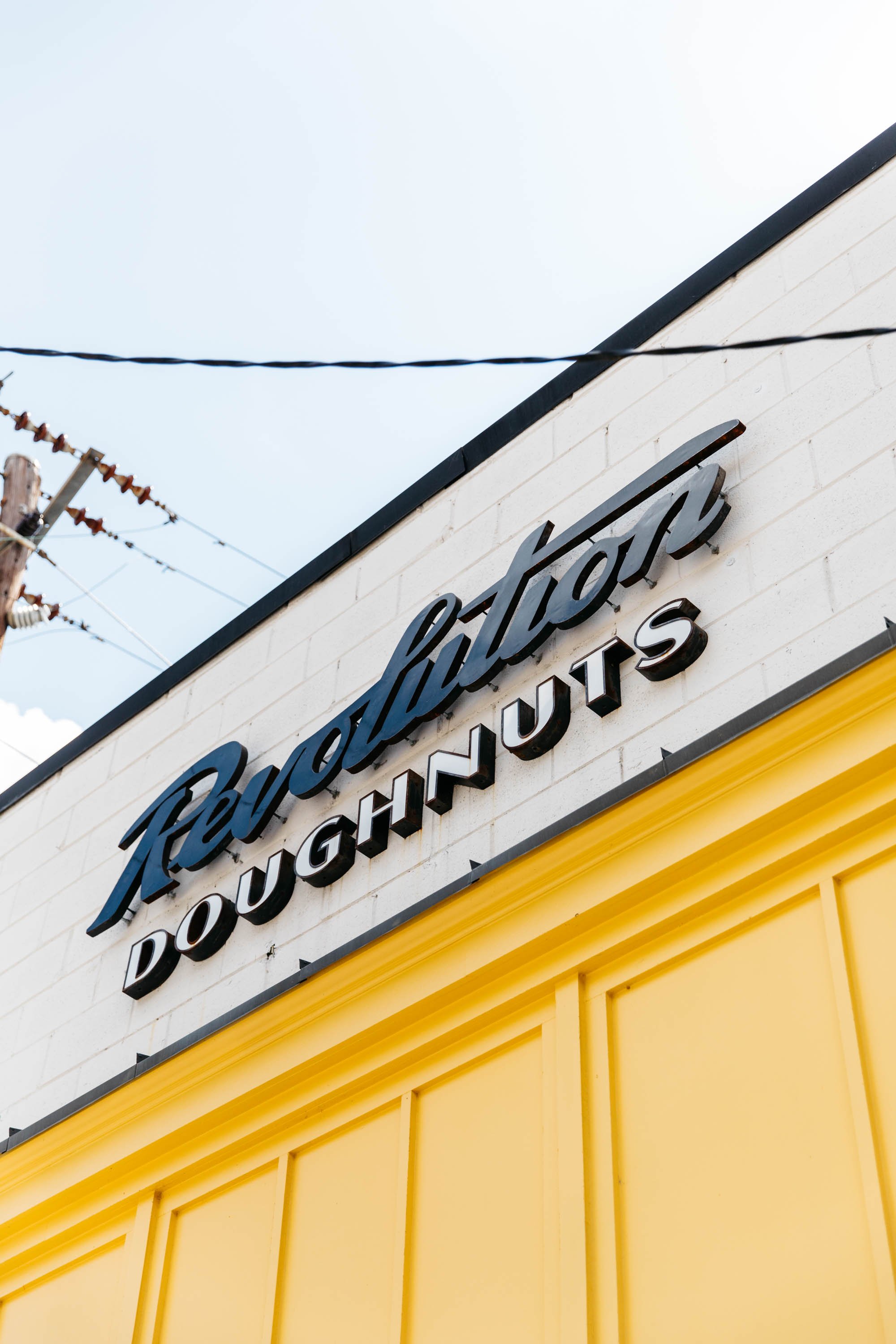 Close-up of a sign on a building that reads "Fabulous Doughnuts" with a bright yellow exterior, and power lines and a clear sky in the background.
