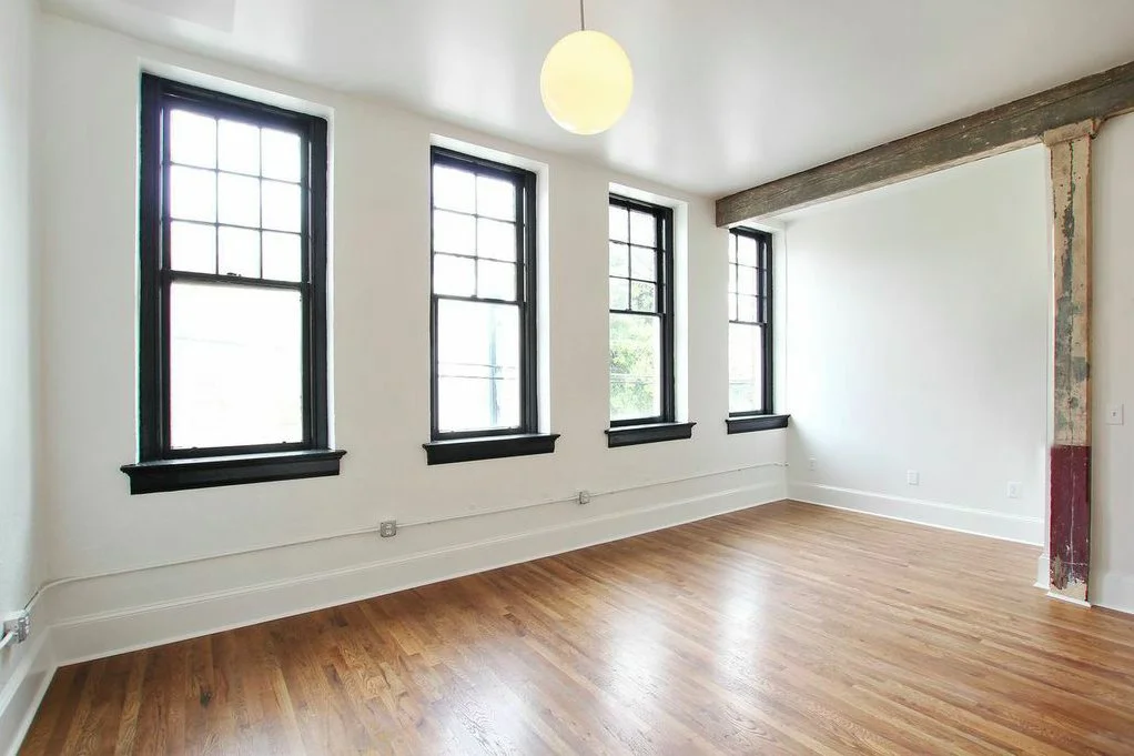 Empty room with four tall black-framed windows, white walls, and hoses running along the baseboard. A round ceiling light hangs from the ceiling, and part of a wooden beam and a partially exposed wall are visible.