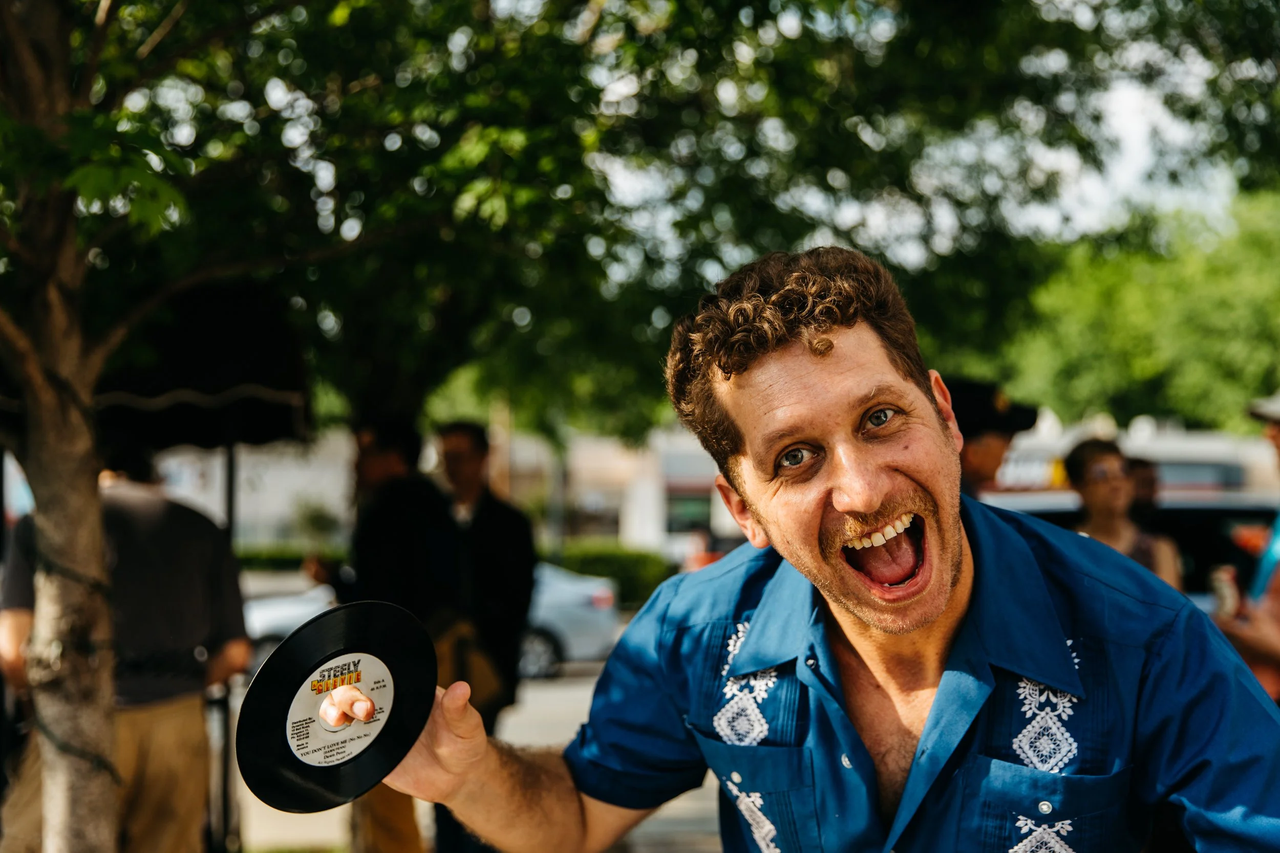A man smiling and holding a vinyl record at an outdoor event with trees and other people in the background.