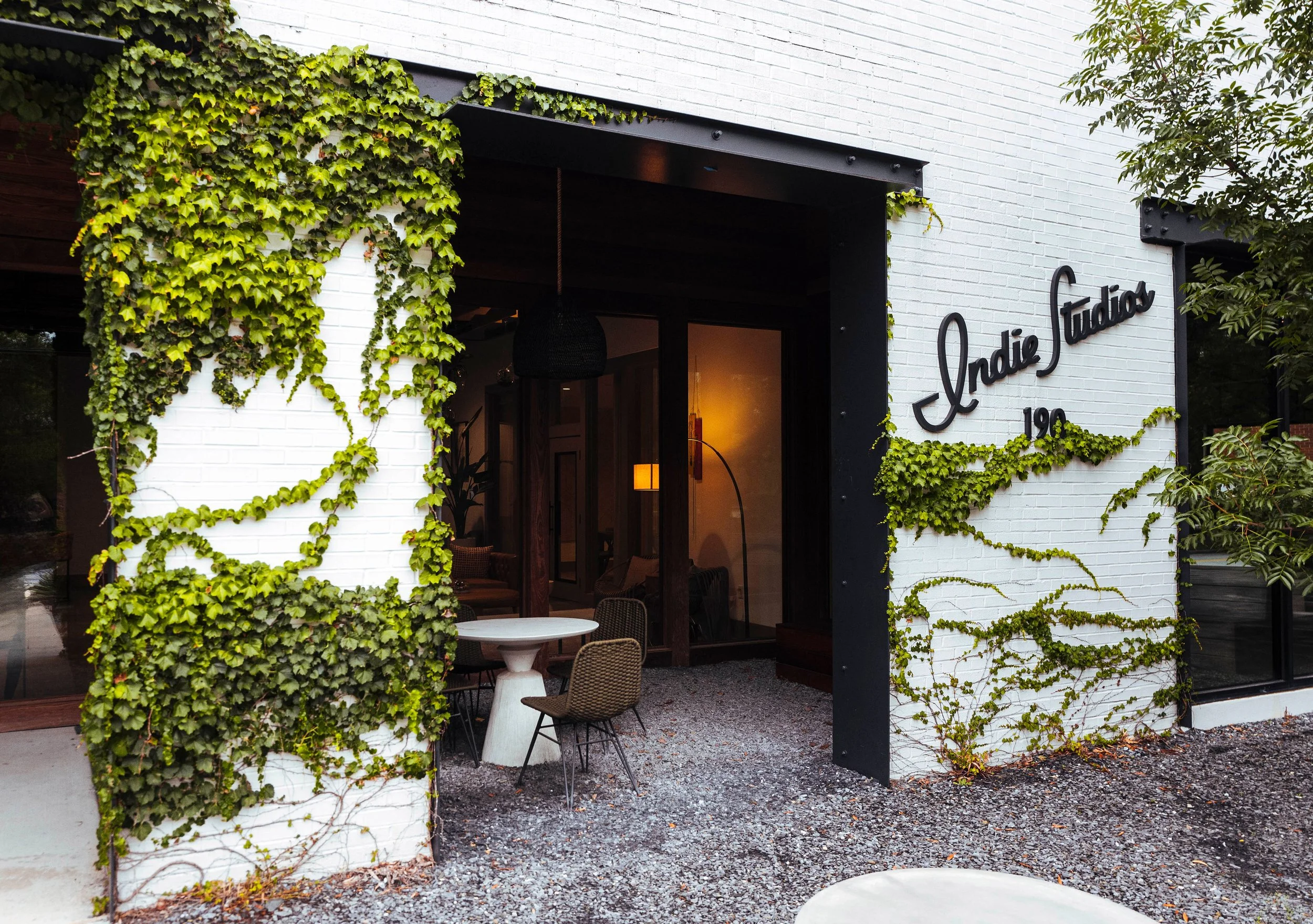 Exterior of a building with white brick walls, black trim, and green ivy climbing the walls. The sign reads 'Crodie Studio' with the number '192' beneath it. Visible inside through the open door are warm lights and interior decor. Outdoor seating inc
