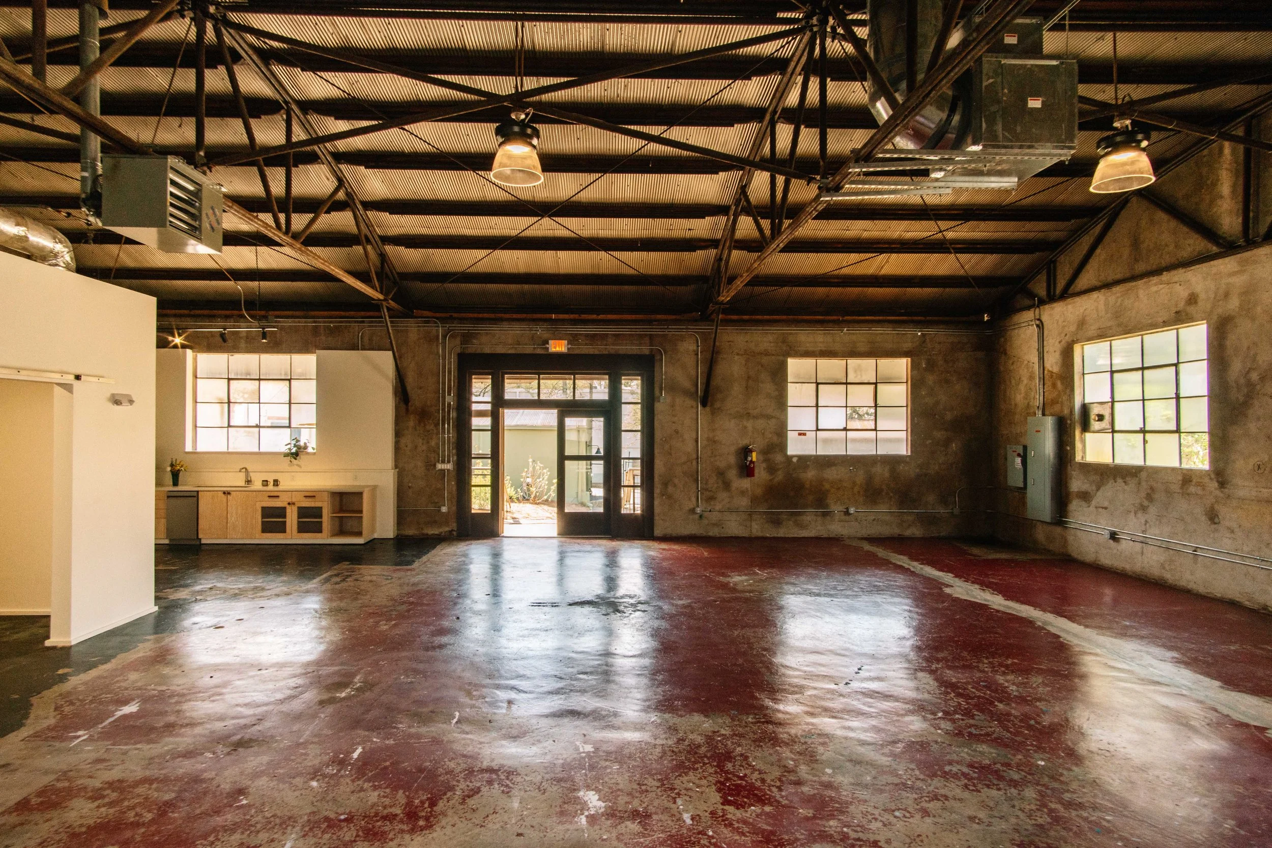 An empty industrial-style room with unfinished walls, exposed ceiling pipes, large windows, and a worn red and black floor.