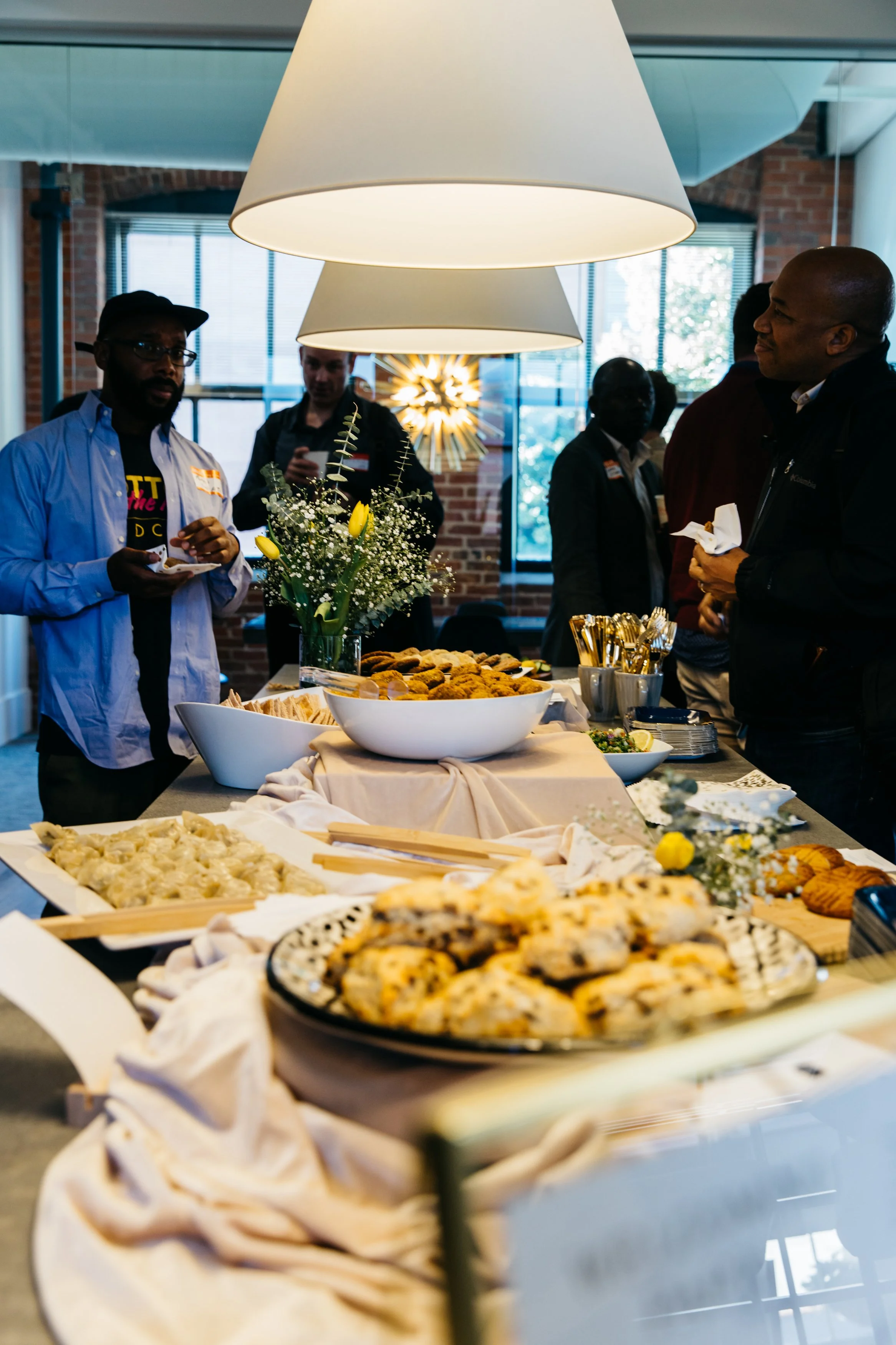 People gathered in a room with large hanging lamps, enjoying a buffet-style meal with various dishes, flowers, and bread on display.