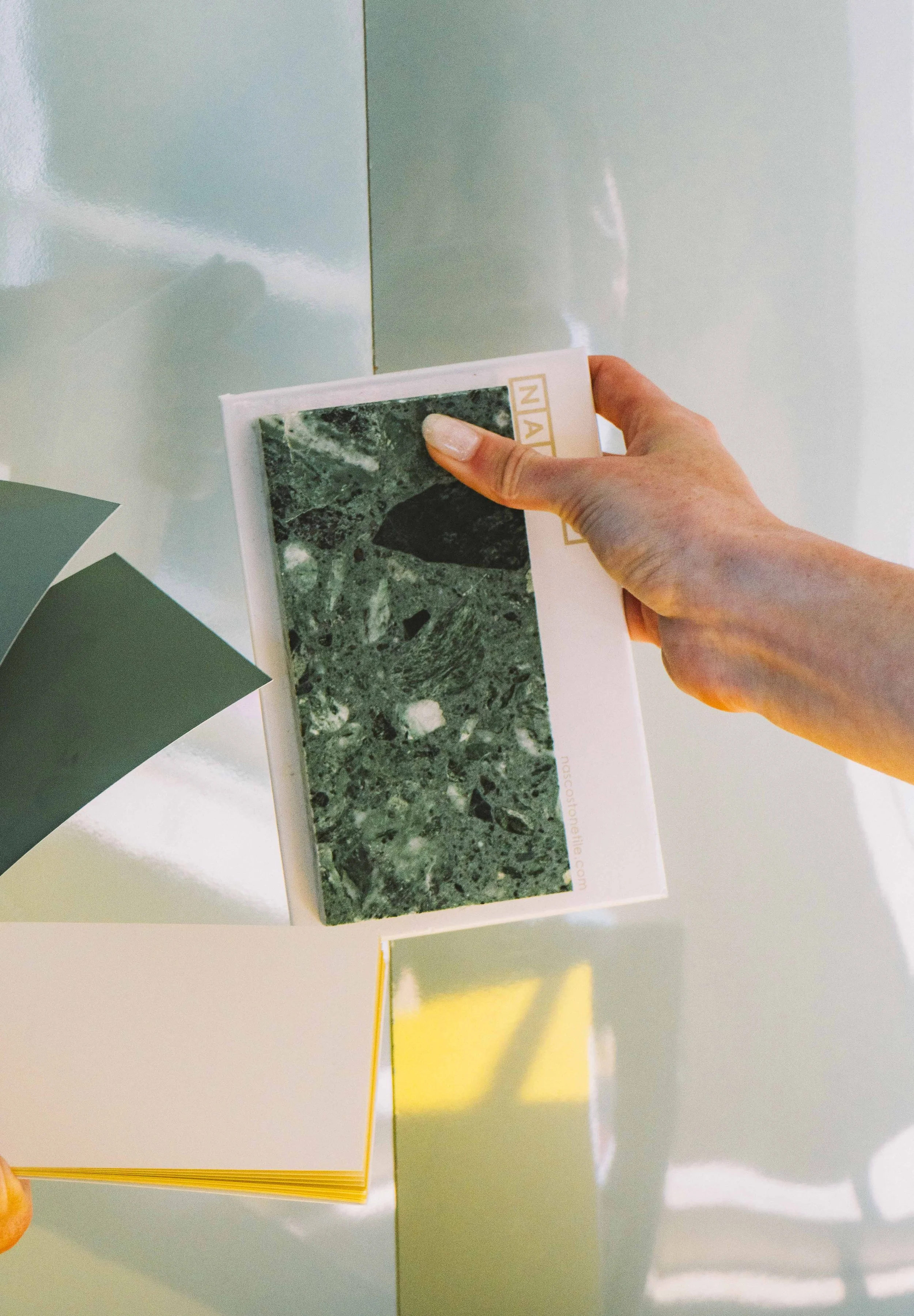 Close-up of a hand holding a brochure with a green marble texture and the text 'NAT' visible on the cover, against a background of pastel-colored walls.