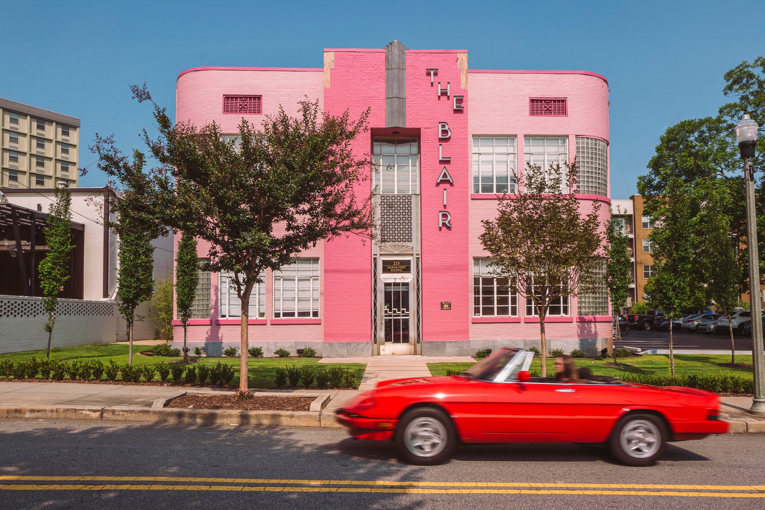 A pink Art Deco building with the words 'The Blair' vertically on the front, surrounded by trees and a sidewalk in front. A red sports car is driving past on the street in the foreground.