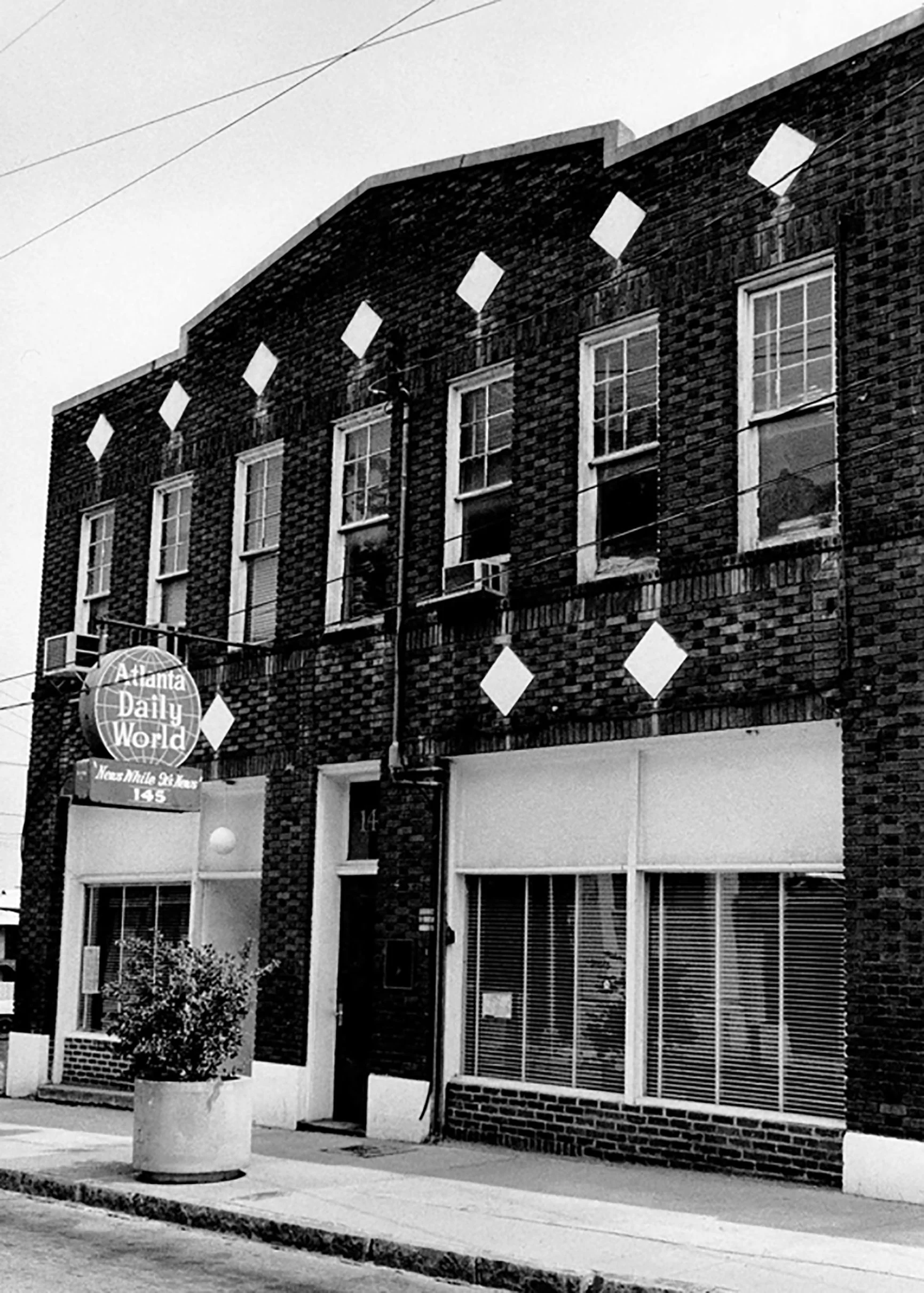 Black and white photo of a brick building with several windows and a sign reading "Atlanta Daily World" on the corner. There is a plant in a large planter on the sidewalk in front of the building, and some power lines are visible above.