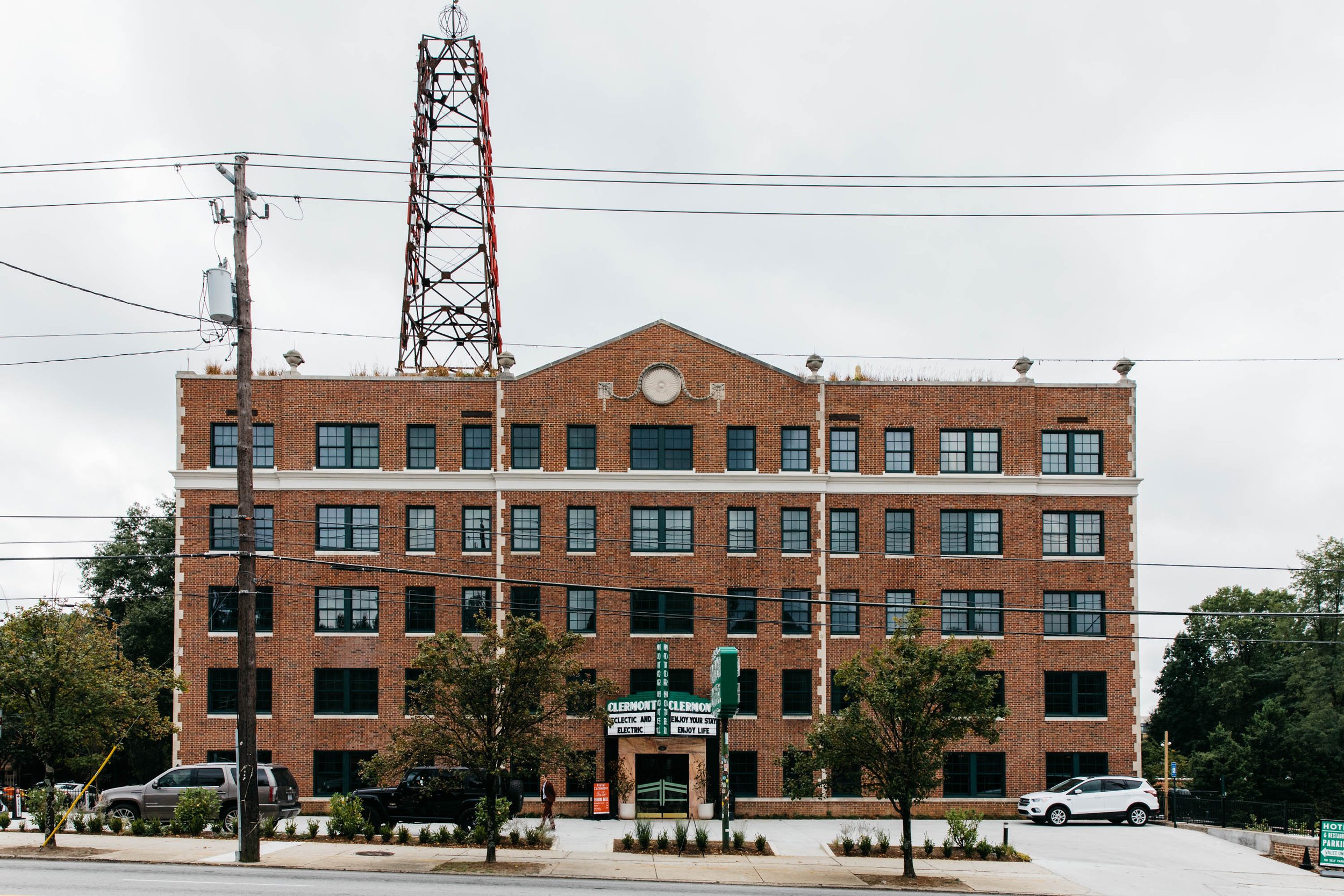 A multi-story brick building with glass windows and a decorative circular emblem at the top center. There are electrical wires and a utility pole in front, with trees, parked cars, and a street view with signs below.