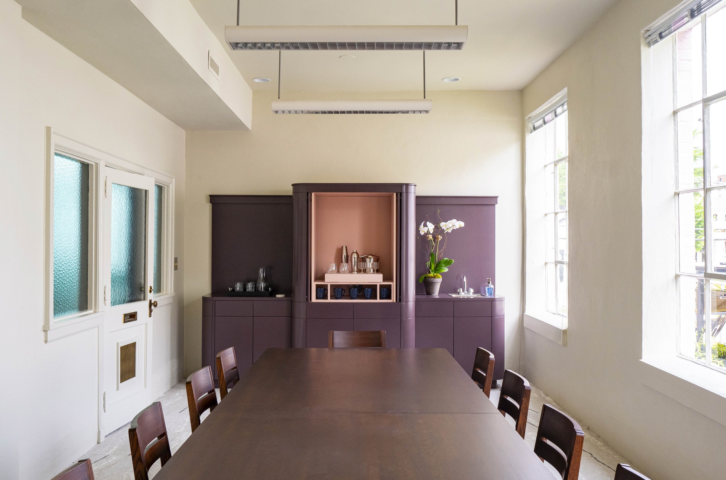 A room with a large wooden table surrounded by chairs, purple cabinetry on the far wall with decorative objects and a plant, large windows on one side allowing natural light, and ceiling lights.