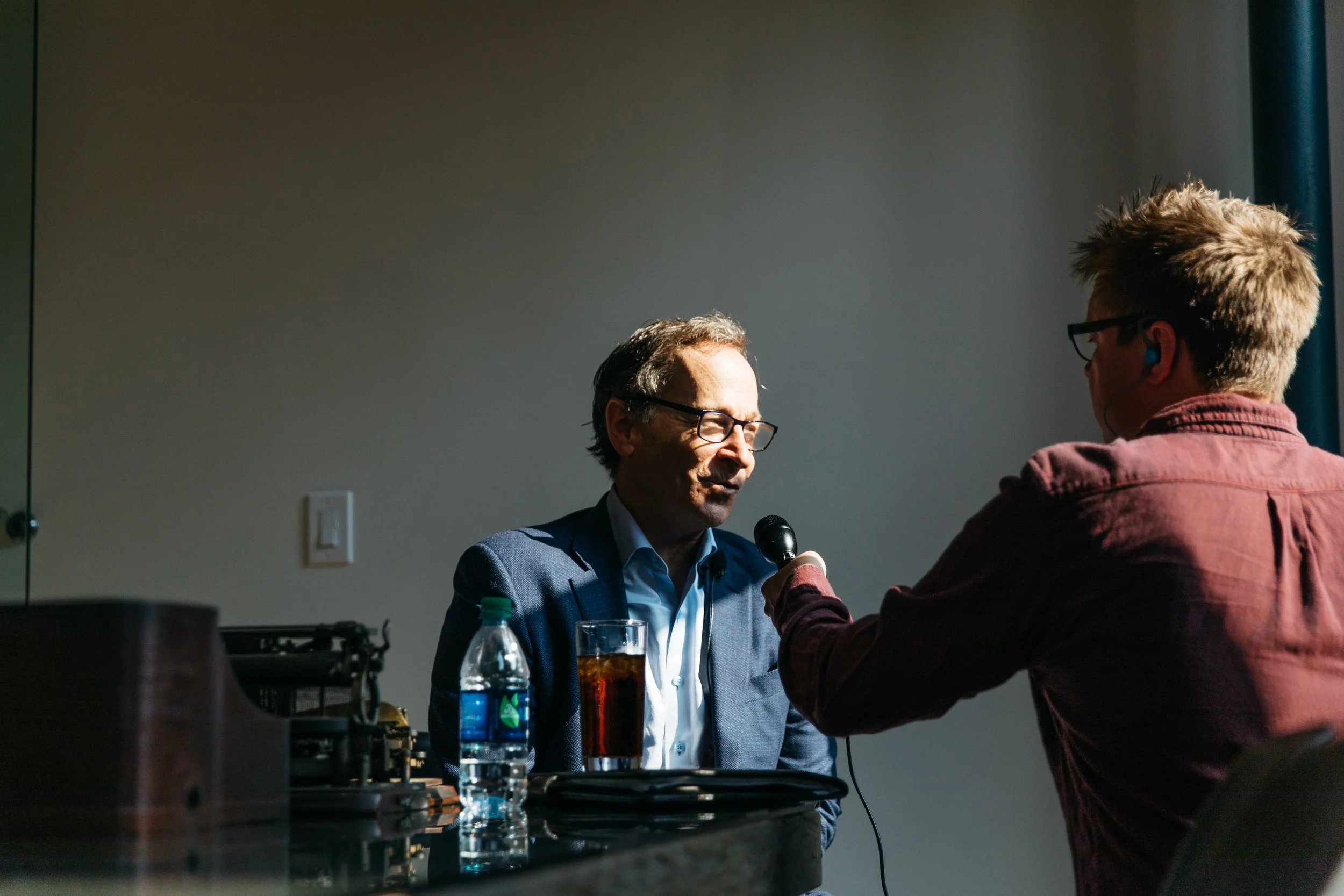 A man in a suit and glasses is seated at a table, speaking into a microphone held by a young woman. The woman has short blonde hair and is wearing a burgundy jacket. There are bottles, glasses, and equipment on the table, with a plain wall and a ligh