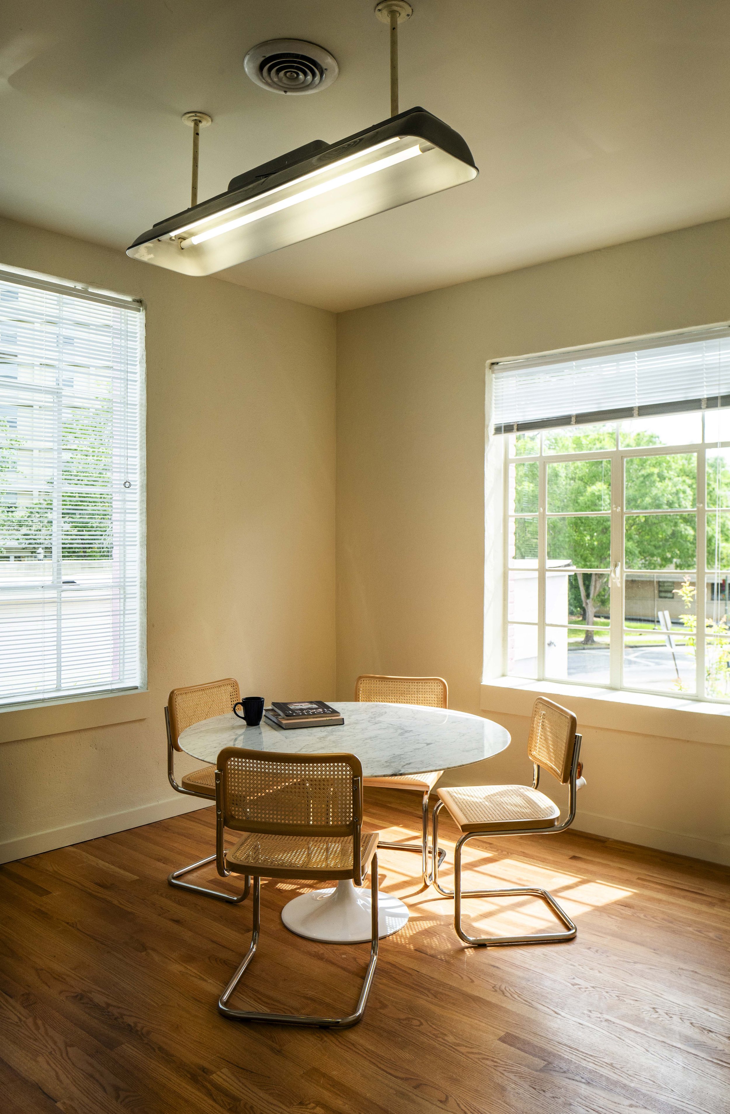 A small dining area with a round marble-top table, four tan woven chairs, a black coffee mug, and a book, next to large windows with blinds letting in natural light, and wooden flooring.