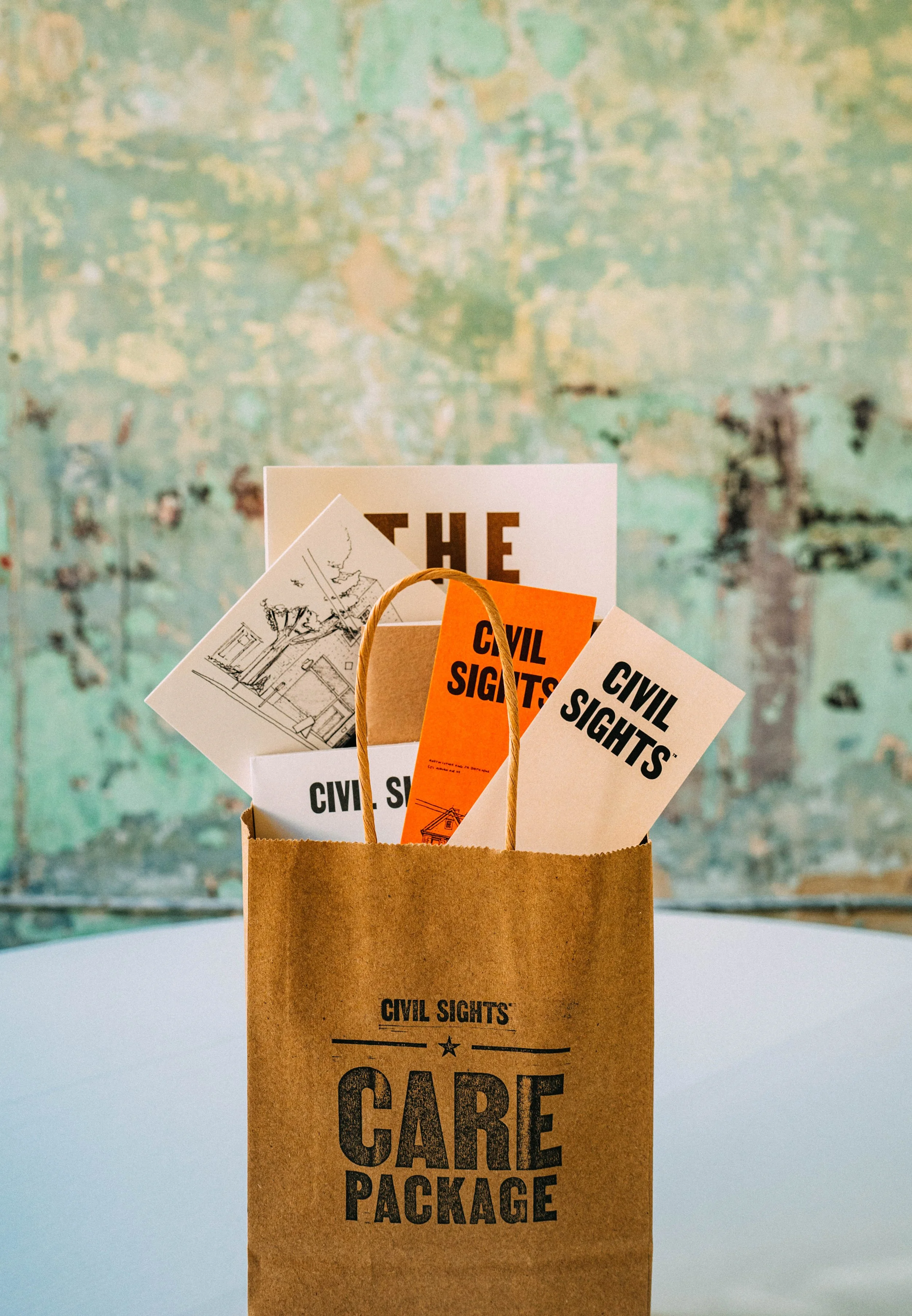 A brown paper care package bag with several civil sight cards sticking out, set against a distressed green and blue wall.
