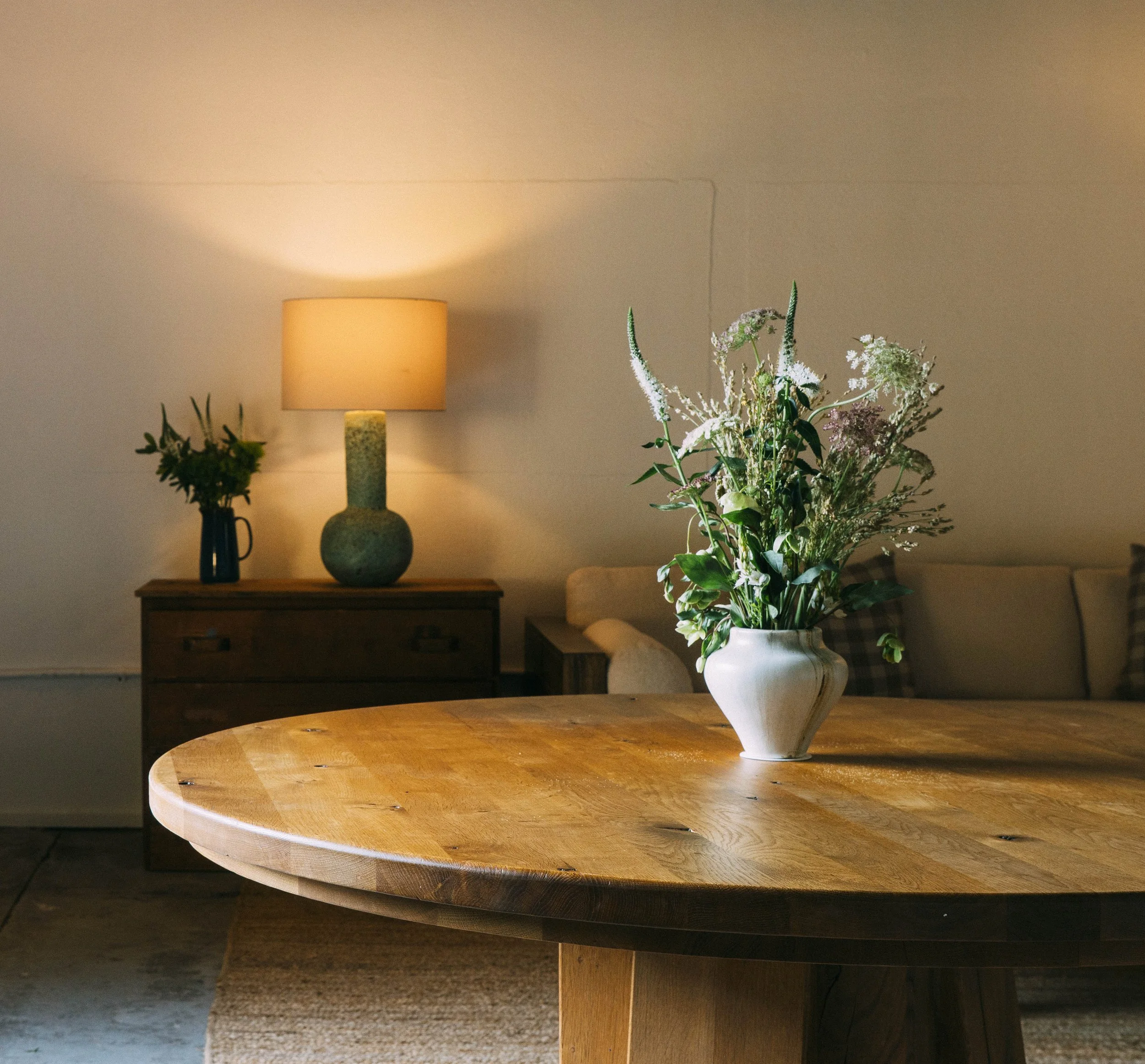 A large round wooden table with a natural finish, and a white ceramic vase filled with tall mixed flowers. In the background, there is a wooden nightstand with a tall green ceramic table lamp and a small dark-colored vase with greenery. The wall behind has a beige color with visible seam lines.