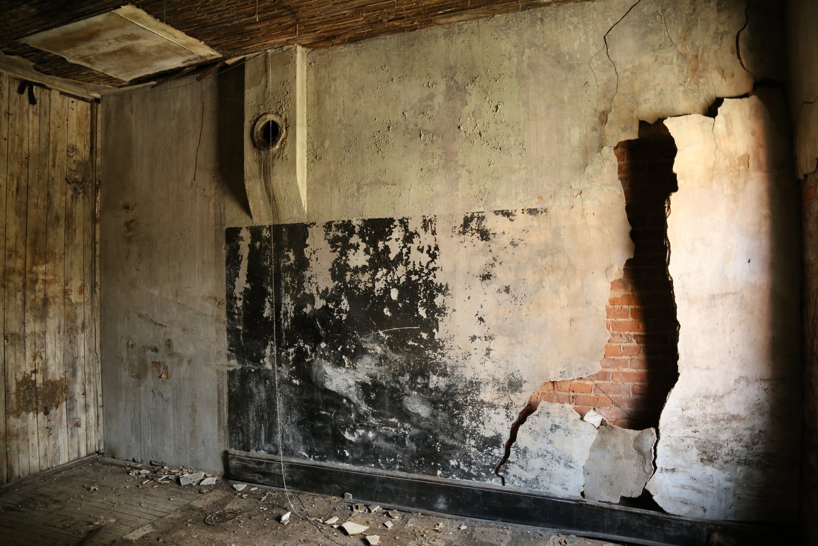 A damaged room with a cracked wall, missing plaster, exposed brick, and debris on the floor.