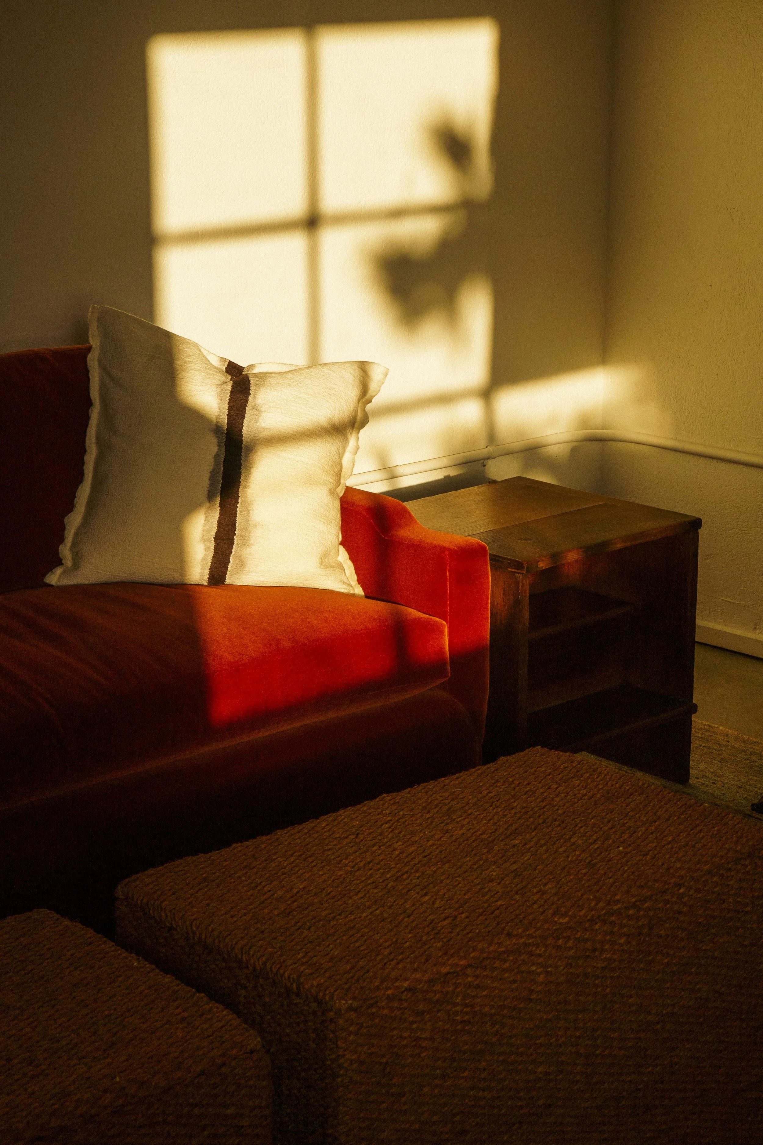 A living room corner with sunlight casting window grid shadows on the wall. A beige pillow with a dark stripe rests on a reddish-orange sofa next to a dark wooden side table. The floor features a textured brown rug.