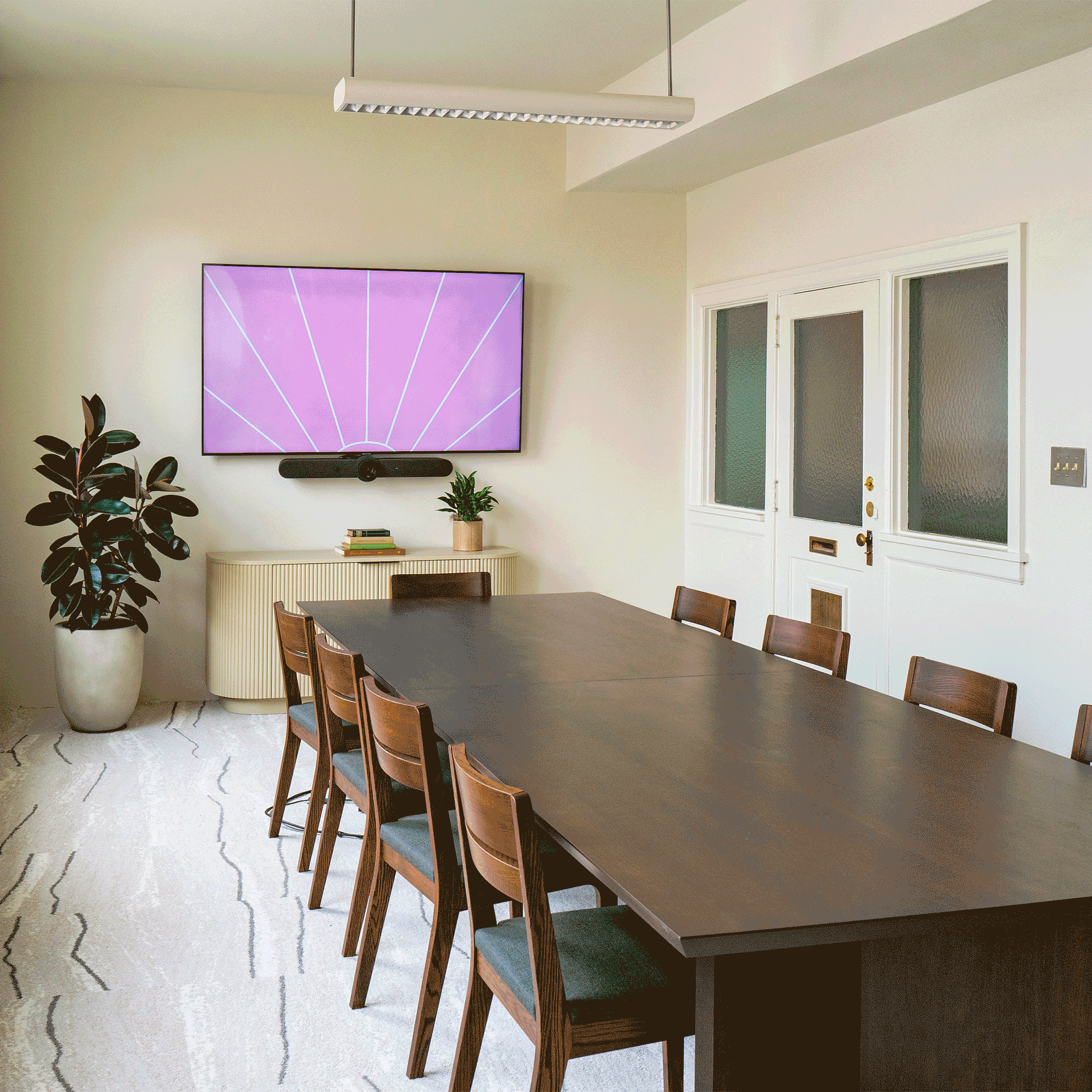 A modern living or dining room with a long dark wooden table and six matching chairs. A large flat-screen TV displaying a purple geometric pattern is mounted on the wall. Below the TV is a soundbar and some books. To the left, there is a tall, large potted plant with dark green or blackish leaves. To the right, there are some white framed windows and glass doors with white trim, one of which has a gold doorknob. The room has clean, minimalistic decor with light-colored walls and a patterned light carpet on the floor.