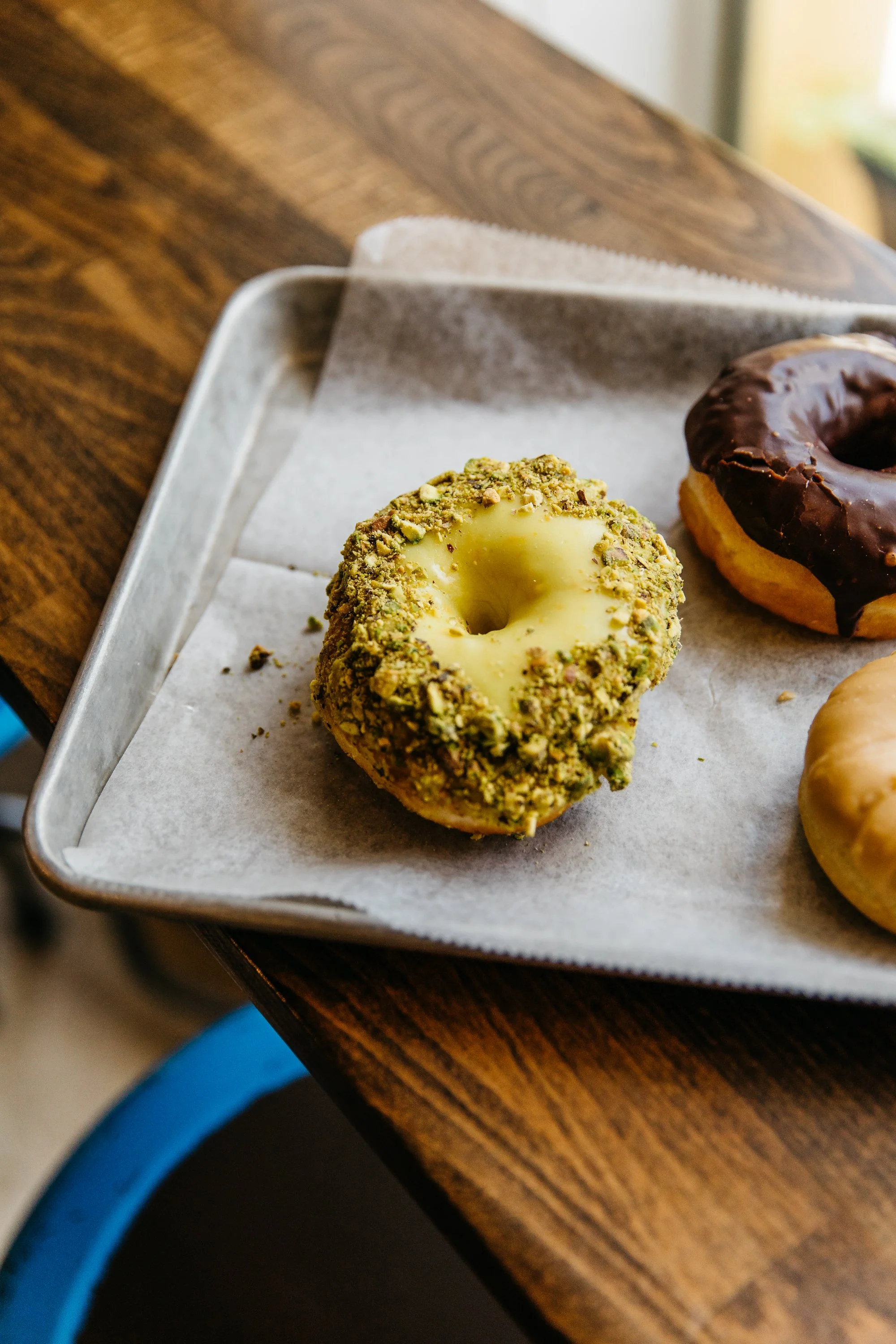A close-up of a tray with assorted donuts, including one topped with crushed pistachios and white icing, and another dipped in chocolate glaze.