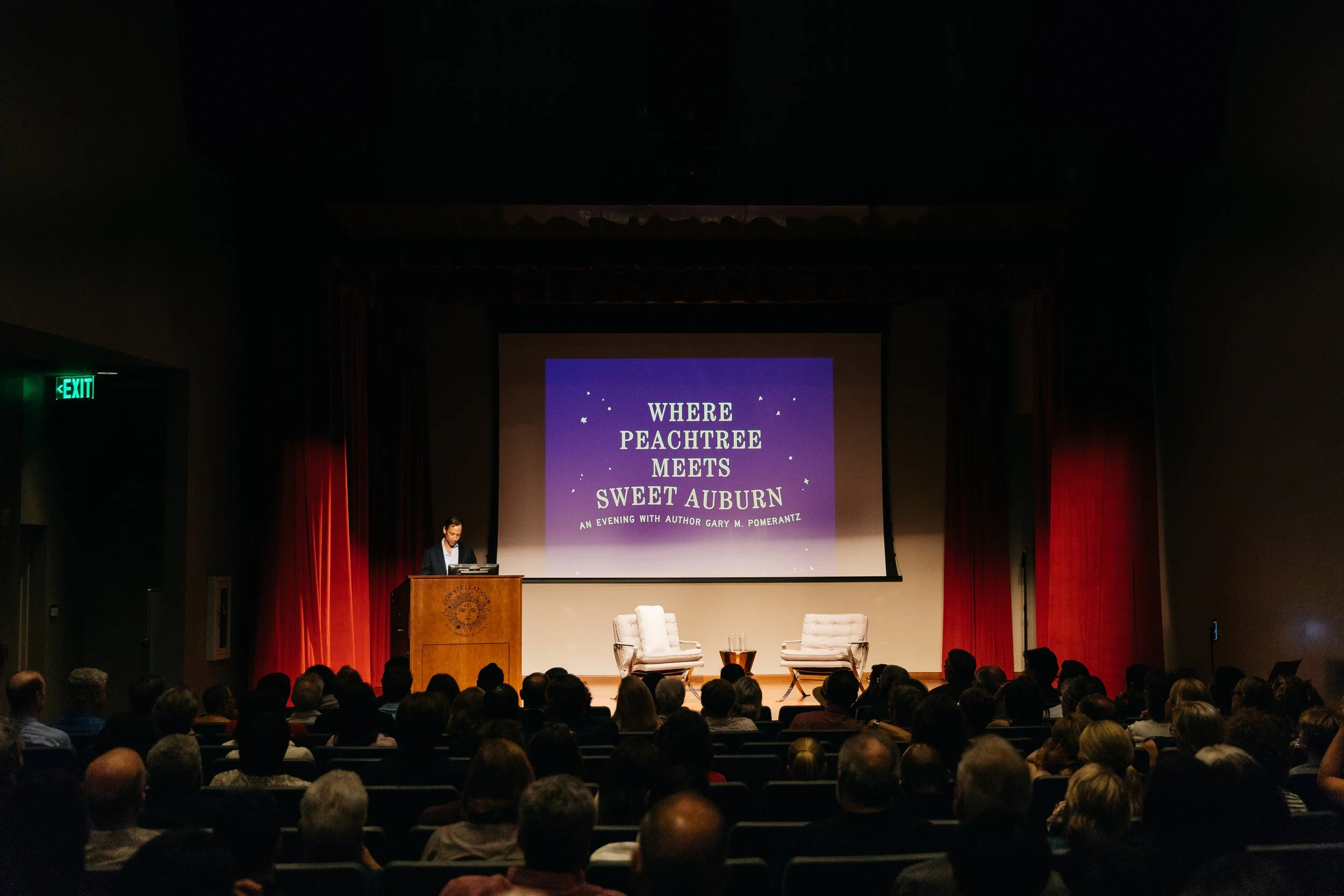 A presentation event titled 'Where Peachtree Meets Sweet Auburn' with an author speaking at a podium in a large theater filled with seated audience members. The screen behind displays the event details.