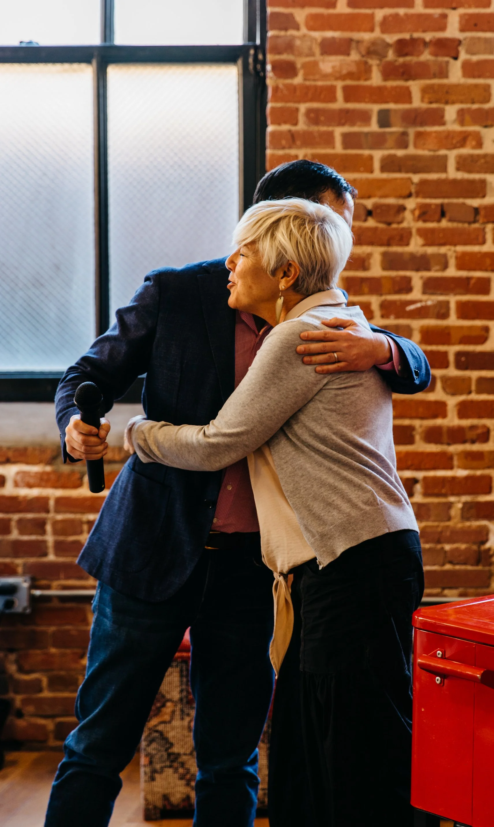 A man and a woman hugging in an indoor setting with brick walls and a window in the background. The man is holding a microphone and is partially visible. The woman has short blonde hair and is wearing a beige sweater and earrings.