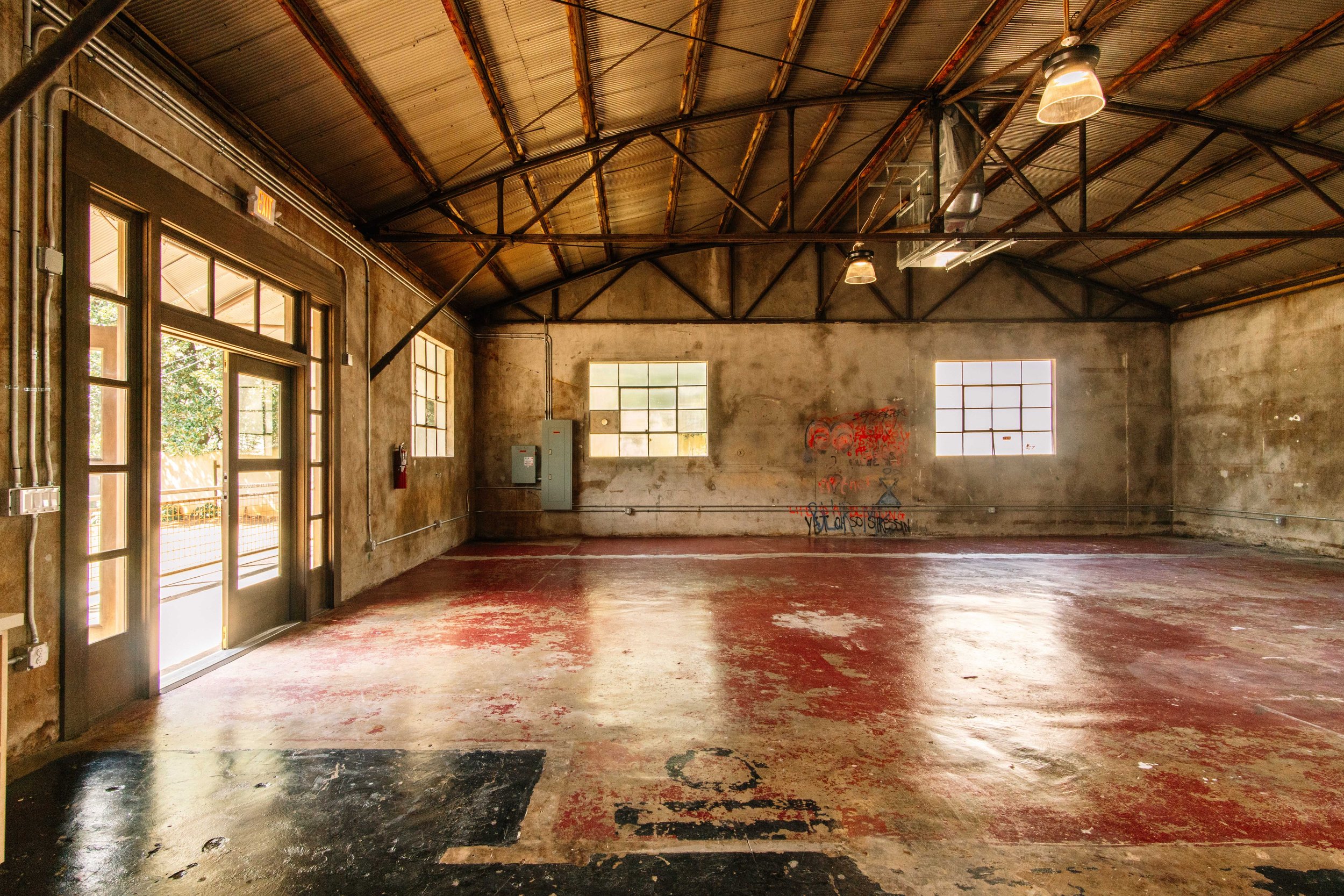 Empty warehouse or studio with large windows, graffiti on wall, and a mixture of concrete and wooden floor.