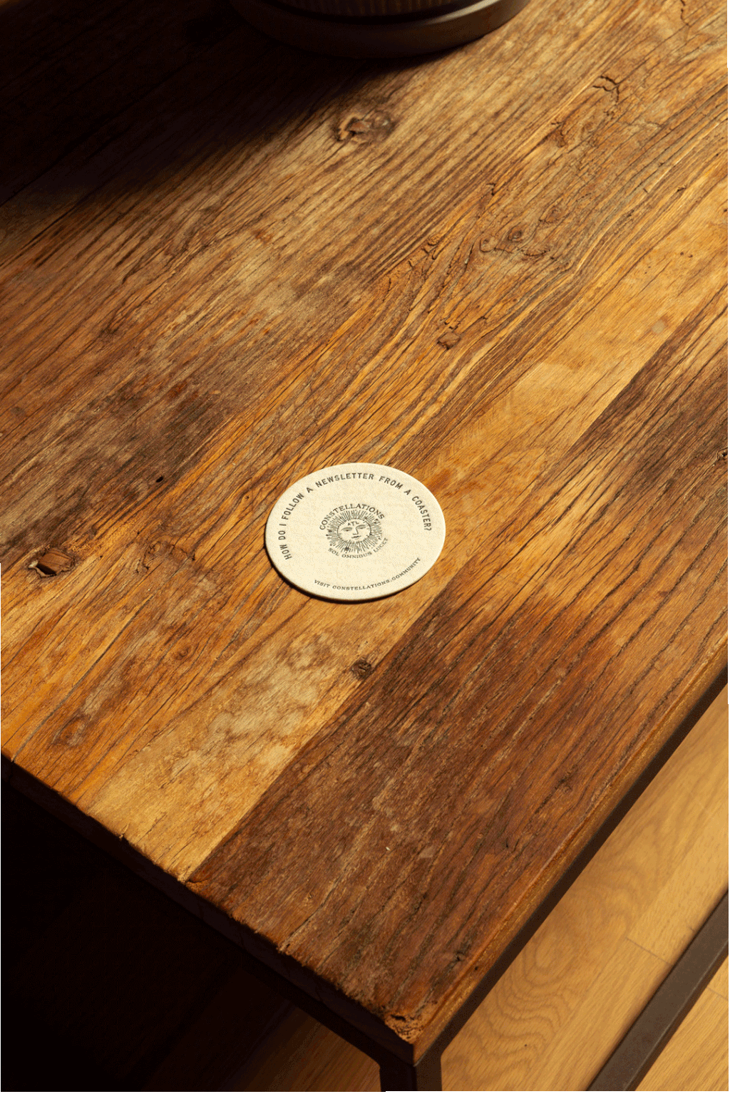 Close-up of a rustic wooden table with a round sticker in the center that says 'How do I follow a newsletter from a coaster?' and features a sun illustration and the website 'constellations.com/subscribe'.