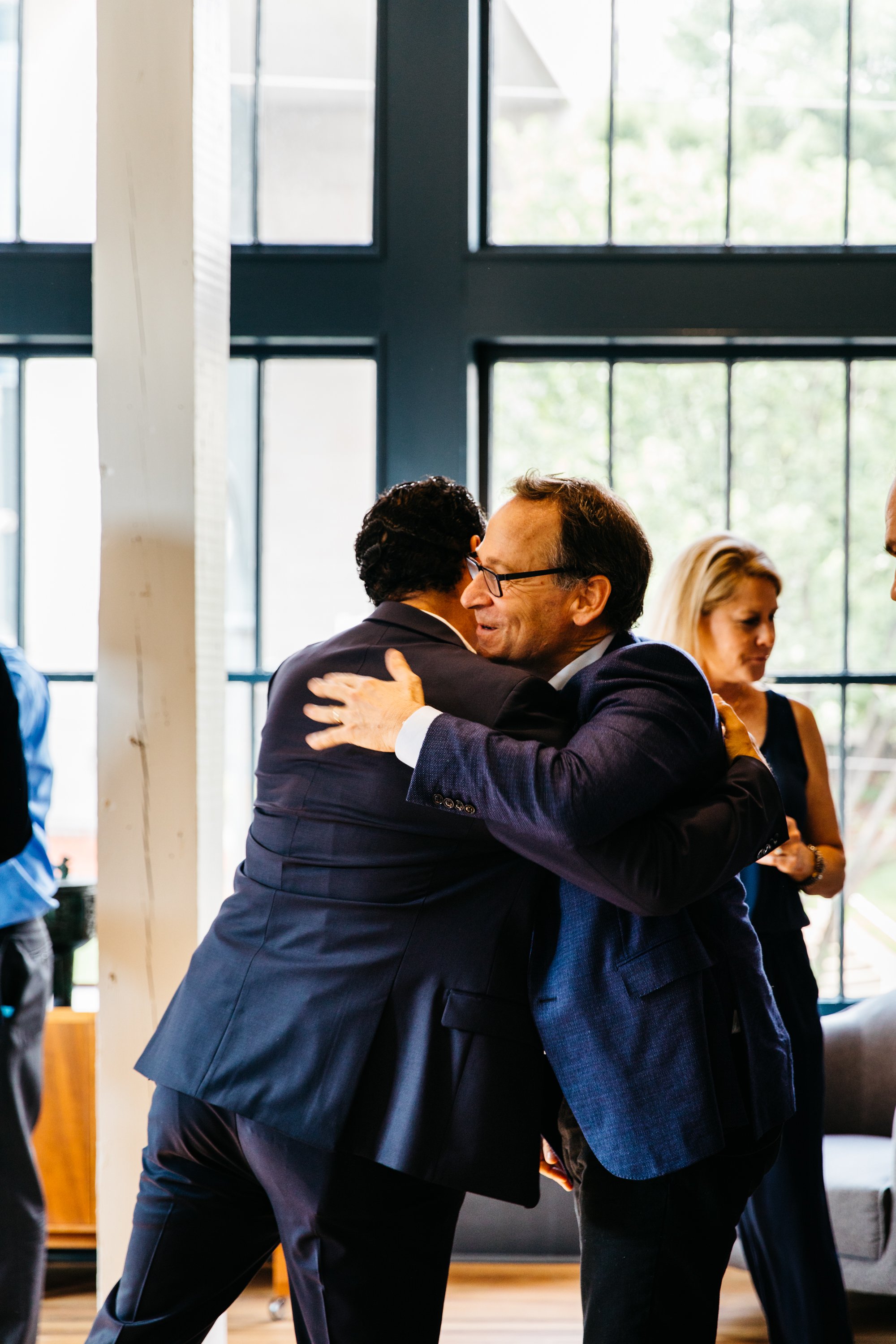 Two men in suits hugging at an indoor event, with large windows and other people in the background.