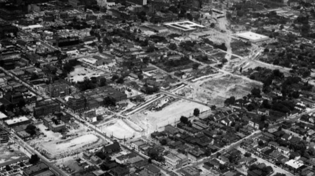 Aerial black and white photo of a city with streets, buildings, and open areas under development or construction.