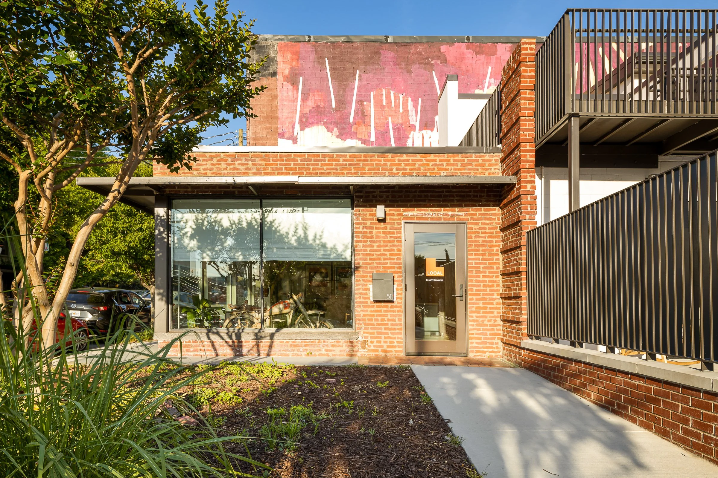 A modern brick building with large glass windows and a gray door, surrounded by greenery, trees, and parking cars in the background.