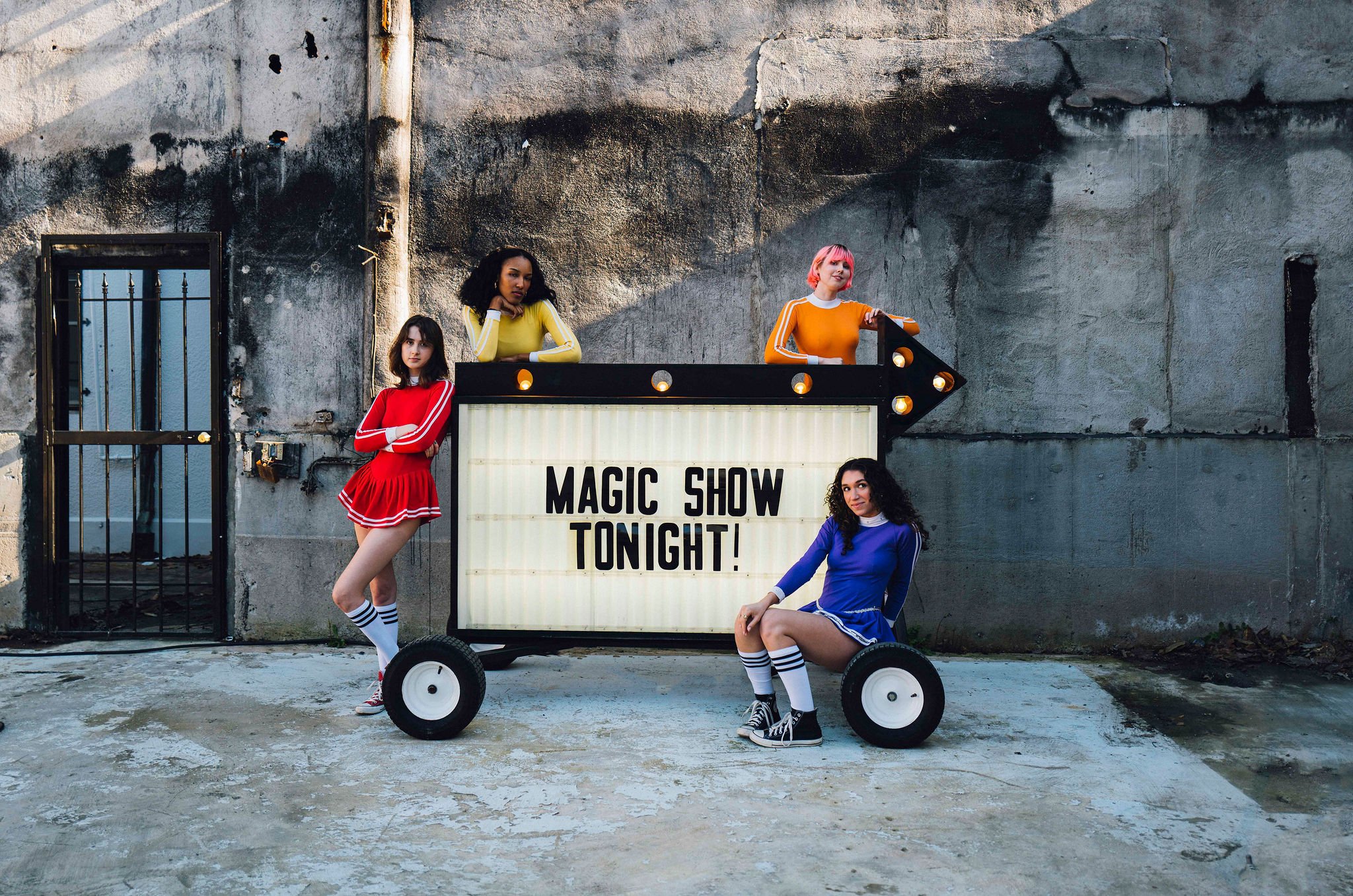 Four young women in colorful athletic outfits posing outside next to a mobile sign that reads 'Magic Show Tonight!,' against a worn concrete wall with a small barred window.