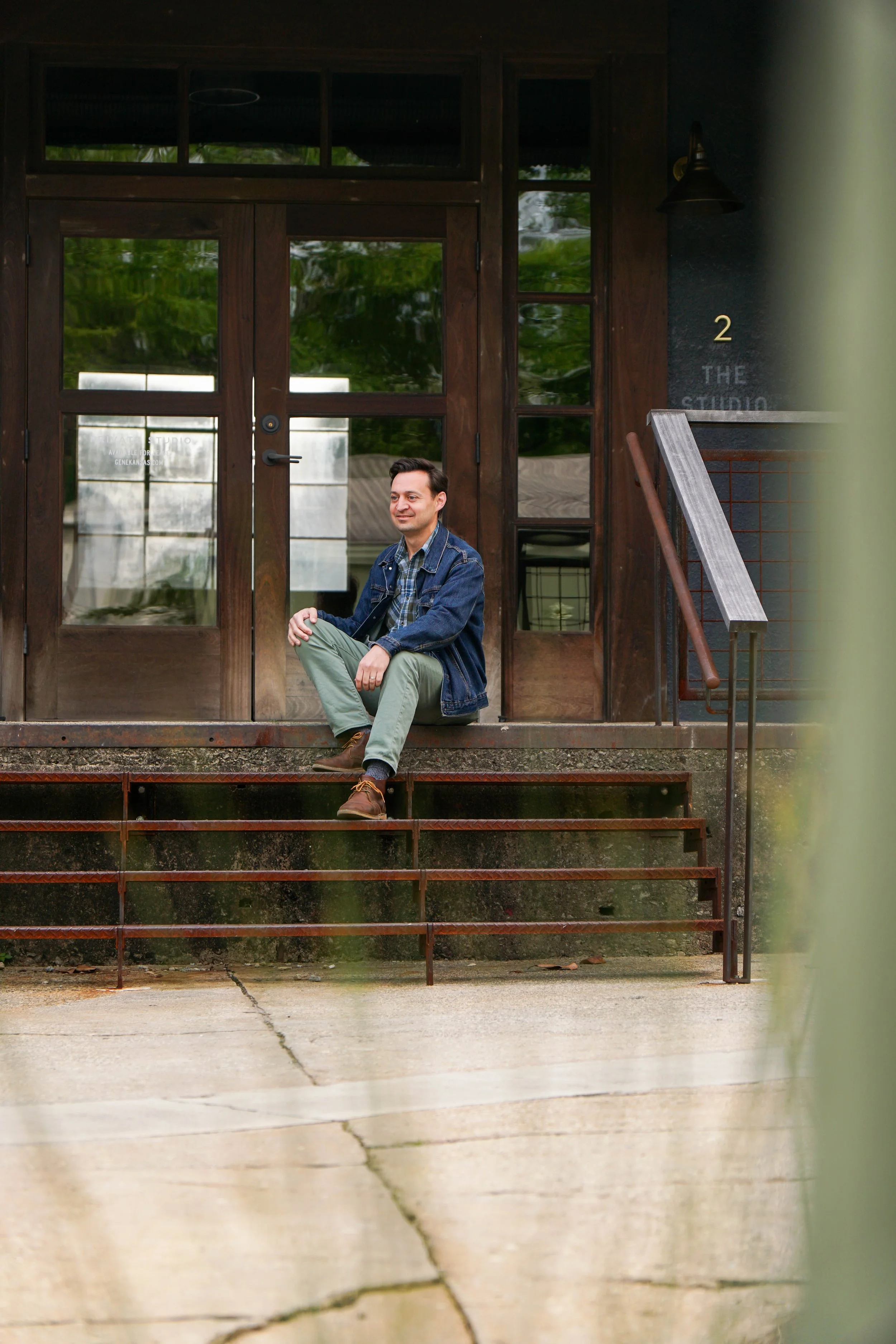 A man in a plaid shirt and blue denim jacket sitting on steps outside a modern building with wooden doors and large windows, with greenery reflected in the glass, and the building has a black exterior wall and a sign with the number '2' and the words 'THE STUDIO' next to the door.