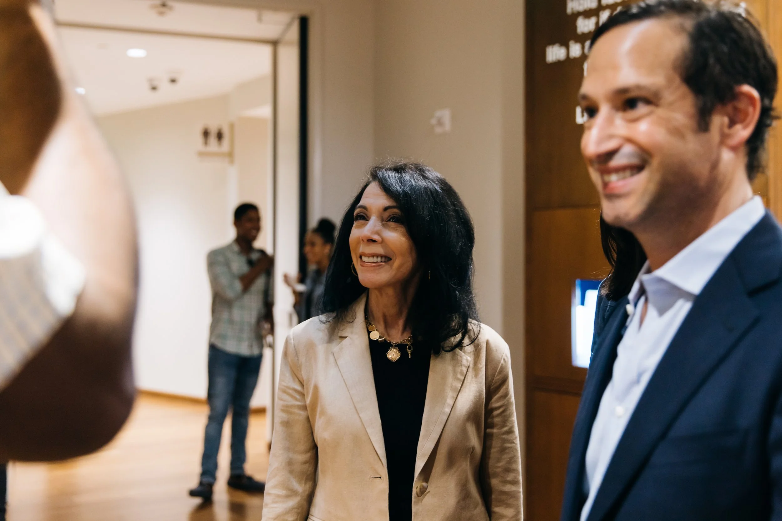 Smiling woman with black hair and a beige blazer talking with a man with dark hair and a blue blazer in a conference or social event setting. Blurred background with other people and signs.