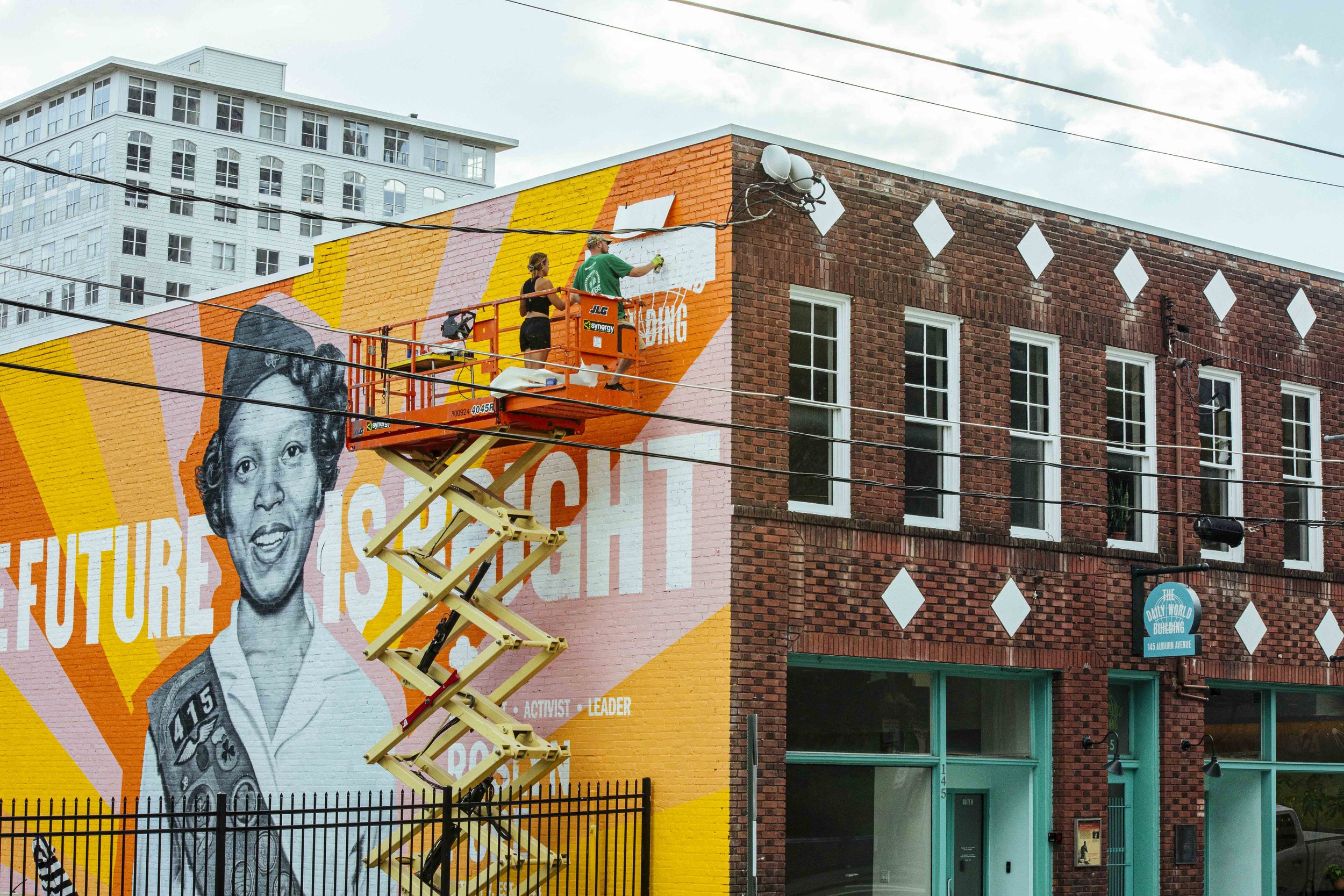Two artists painting a large mural on the side of a brick building, featuring a young girl's portrait with a rainbow background. The mural includes words like 'activist,' 'leader,' and 'storyteller.' One artist is standing and the other is on an elevated lift.