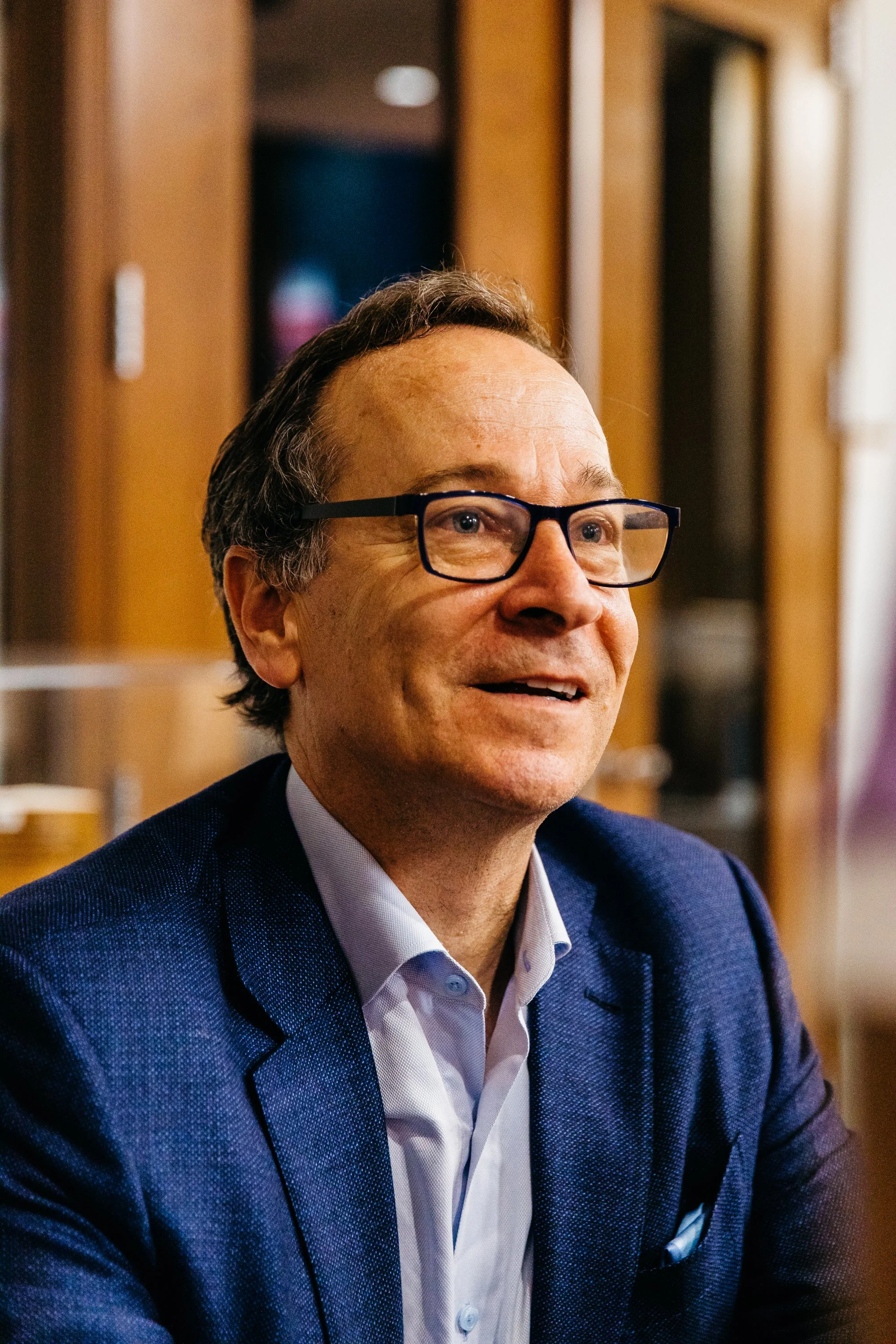 Close-up of a middle-aged man with glasses, wearing a blue suit and white shirt, smiling in an indoor setting.