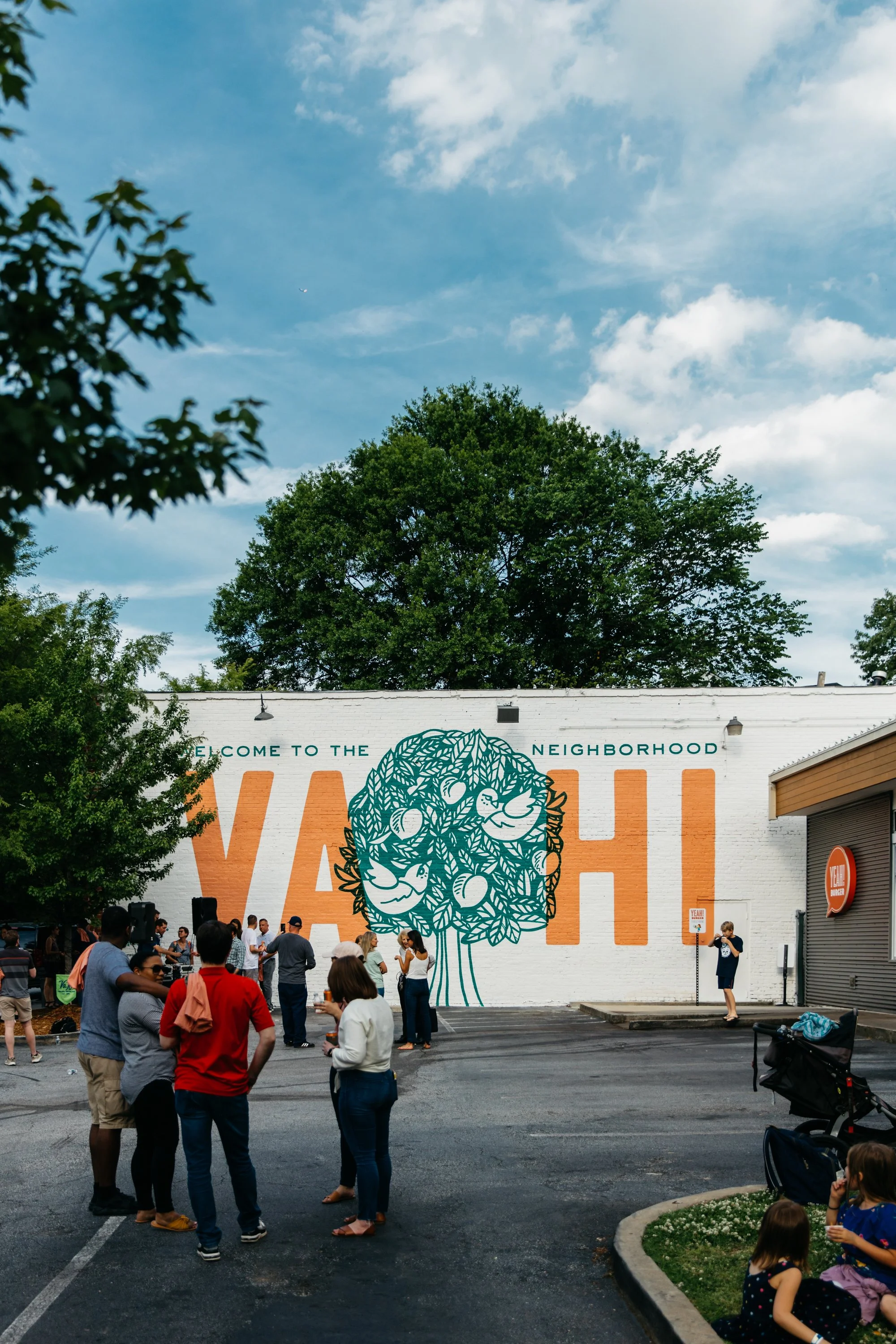 A group of people gathered outdoors in front of a large mural with the words 'Welcome to the Neighborhood' and a stylized tree design, on a sunny day with blue skies and some clouds.