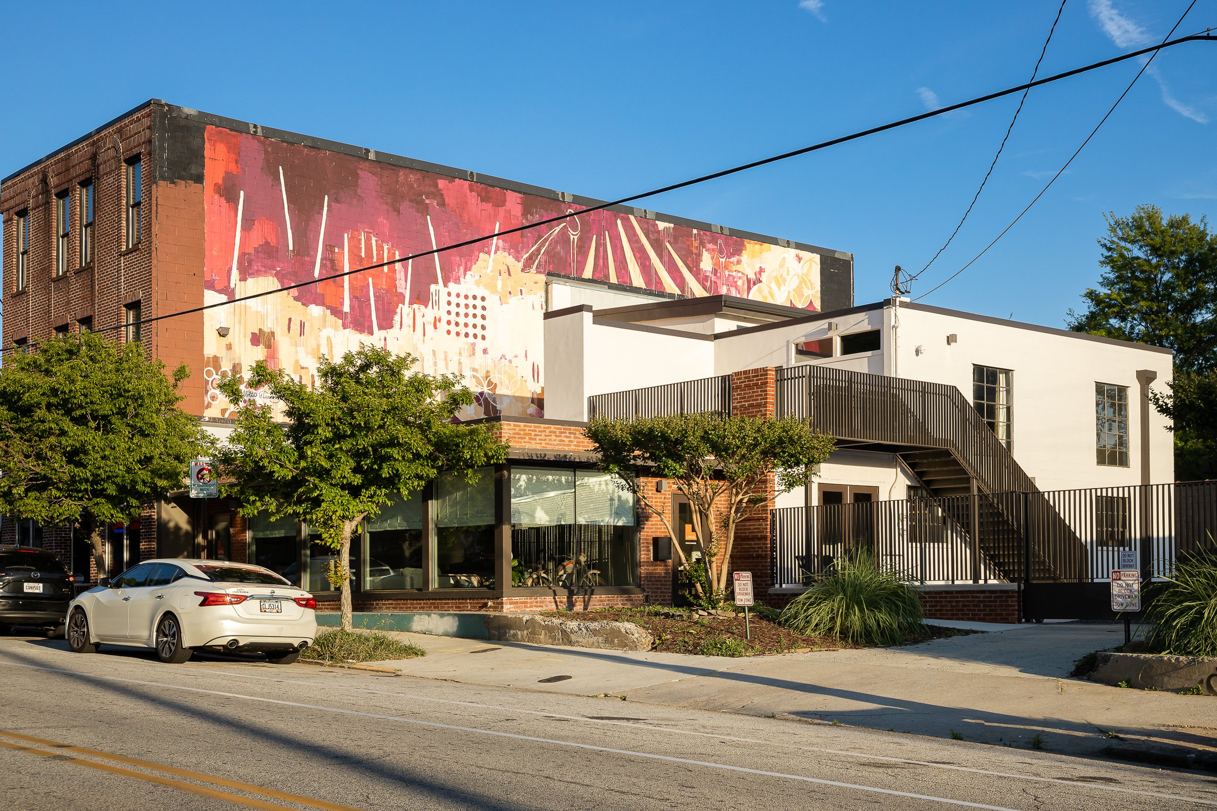 Buildings with a large colorful mural on the upper part of the wall, trees, parked cars, and street signs in the foreground on a sunny day.