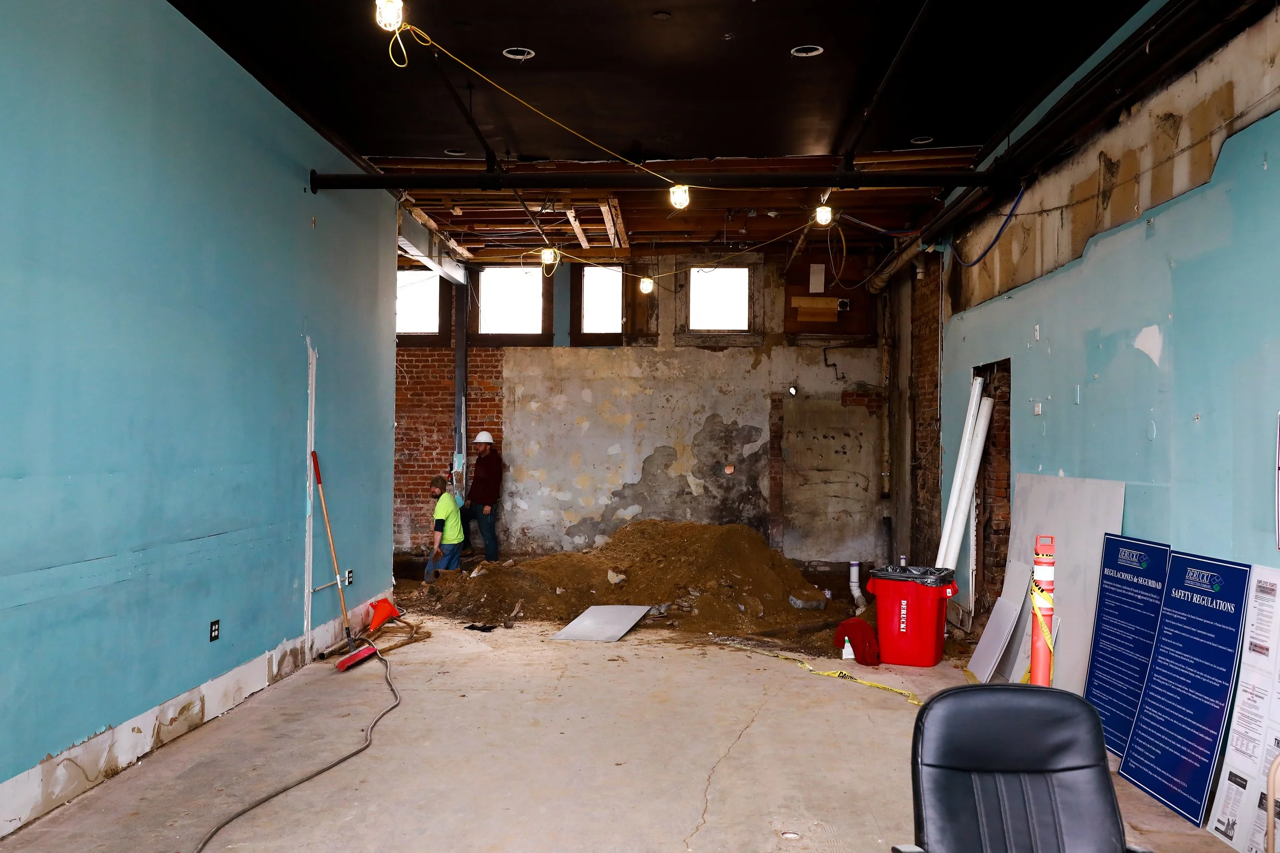 Interior of a building under renovation, with exposed brick and wall damage, construction workers, construction materials, and large windows
