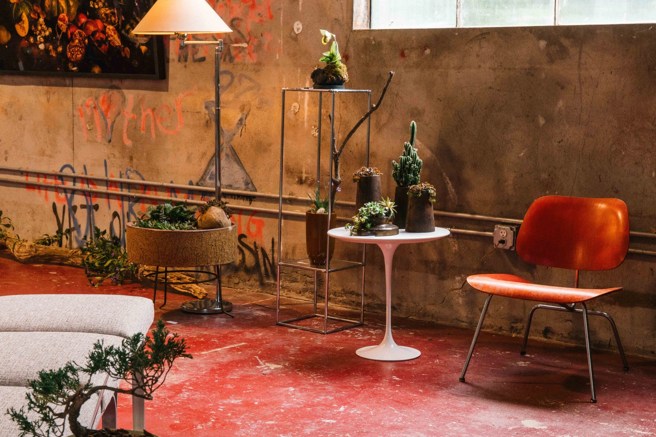 Indoor decor with plants on tiered metal stand, a white round table, orange chair, wall with graffiti, and a framed picture or painting on the wall with a lamp above it.