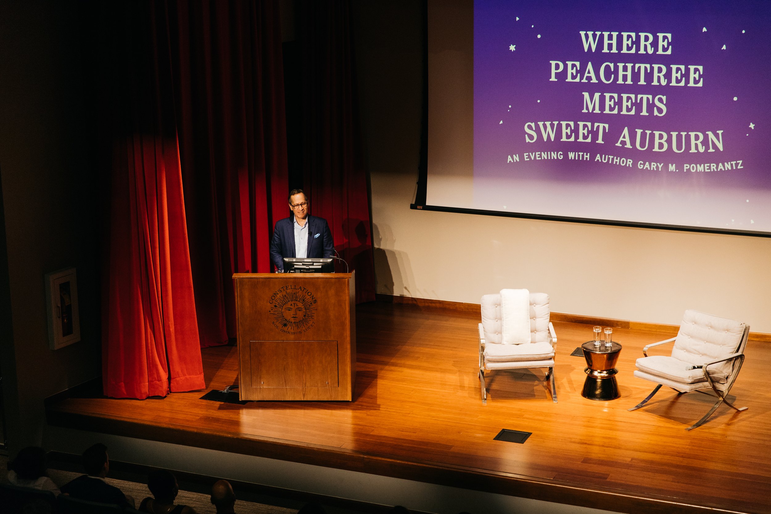 A man at a podium preparing to speak at an event titled "Where Peachtree Meets Sweet Auburn: An Evening with Author Gary M. Pomerantz". The stage features red curtains, two white chairs, and a large purple slide with white text projecting the event's name.