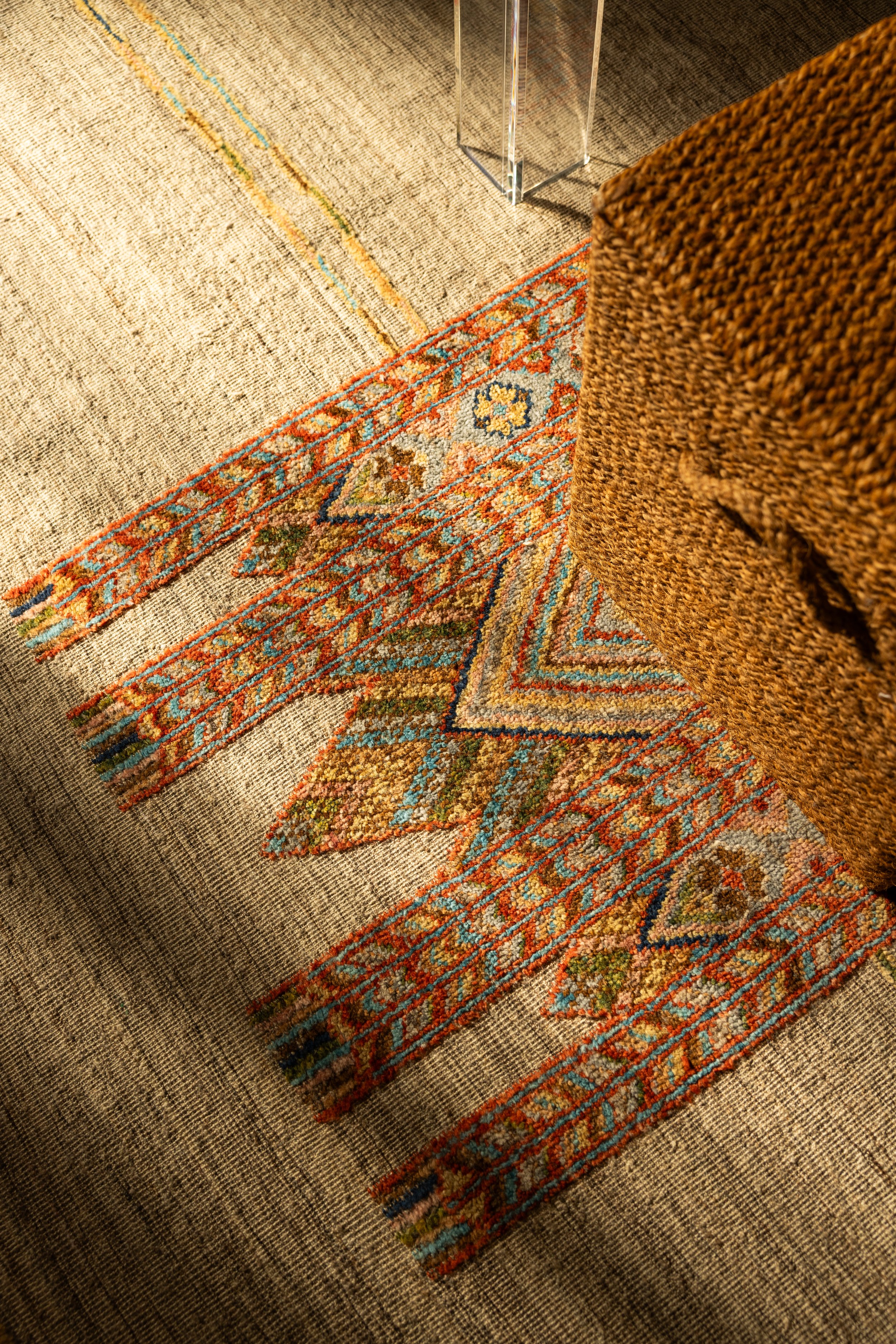 Close-up of a corner of a patterned rug on a beige carpet, with a glass table leg and a woven brown armchair visible.
